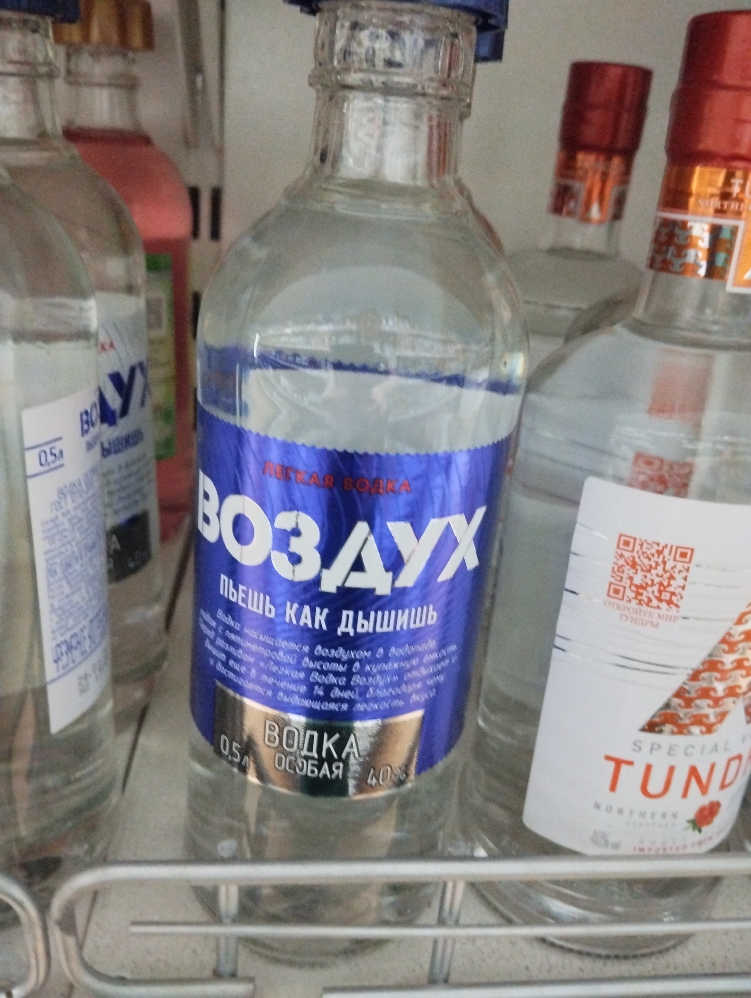 A close-up shot depicts several bottles of vodka displayed on a metal shelf. The central bottle features a blue label with white Cyrillic text, prominently displaying "ВОЗДУХ" and "ПЬЕШЬ КАК ДЫШИШЬ", indicating the brand name "Vozdukh". The label specifies "ЛЕГКАЯ ВОДКА" (light vodka) and details about its production, including being saturated with air in a "vodonade". The bottle is labeled as "0.5л" with a 40% alcohol content.
To the right is a bottle of "TUNDRA" vodka, labeled as "SPECIAL VODKA". A QR code is visible on its label. To the left, part of another vodka bottle is visible, with a label in Cyrillic, including the capacity "0.5л". The location in the city of Krasnogorskoye, Russia, is not explicitly evident from the image itself.