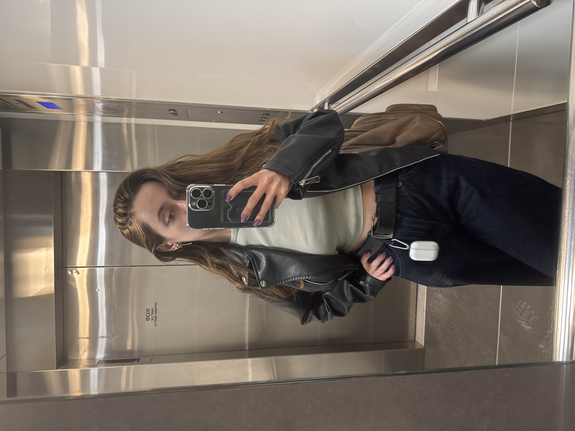 A woman is taking a selfie in an elevator, the interior of which features brushed metal walls and a stainless steel handrail. She is wearing a black leather jacket, a light-colored top, dark jeans with a black belt, and has long brown hair. She holds a phone with a clear case, showing nails painted purple. Attached to her belt are white wireless earbuds. A tan bag is visible behind her on the upper right. The elevator's control panel and other interior elements are reflected in the mirrored surface. The elevator door has Hebrew writing on it, placing this scene in Ramat Gan, Israel.
