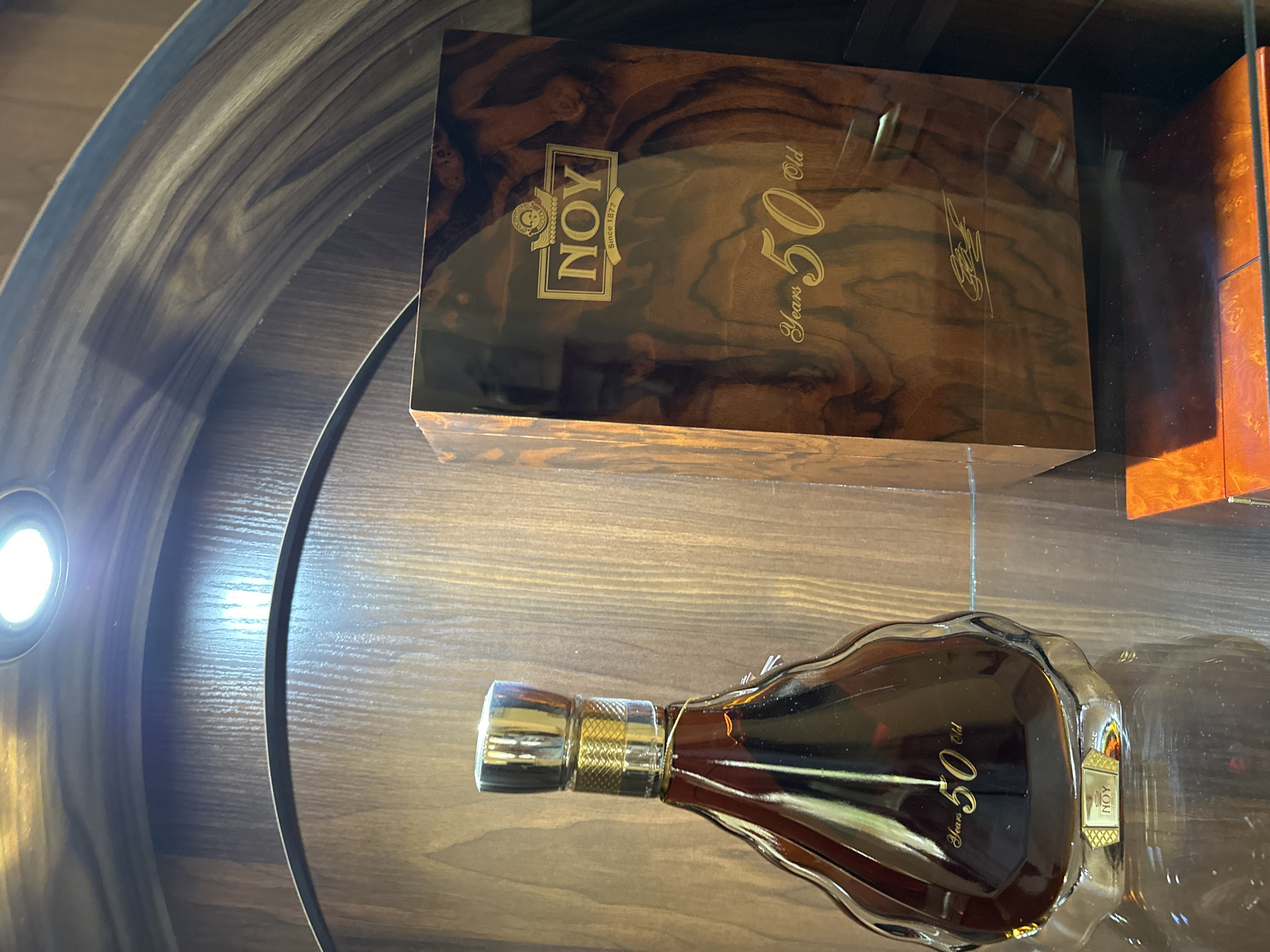 The image displays a high-end bottle of alcoholic beverage and its accompanying wooden box, presented in what appears to be a luxury display setting. The scene is illuminated by an overhead spotlight on the left, creating reflections on the glossy surfaces of the items.
The main subjects are a uniquely shaped glass bottle filled with a dark amber liquid, likely brandy or cognac, and a rectangular, dark wooden box with a rich, glossy finish and prominent wood grain. The bottle features a gold-colored stopper and a small label near its base with the word "NOY." Large golden text "Years 50 Old" is also visible on the body of the bottle, indicating its age.
Behind and slightly above the bottle is the elaborately crafted wooden box. Its lid prominently displays the brand "NOY" in a golden emblem, accompanied by "Since 1877." Below this, "Years 50 Old" is written in elegant golden script, along with an illegible golden signature. The box's surface is highly reflective, showing distortions from the lighting.
The items are situated on a flat wooden surface, part of a larger display unit featuring curved wooden elements, possibly a barrel-like structure or a specially designed shelf. The luxurious presentation and the "NOY" brand, combined with the provided context, suggest that this is an Armenian brandy, likely from Yerevan, Armenia, emphasizing its prestige and maturity. No people are visible, and the activity depicted is the static display of these premium products.