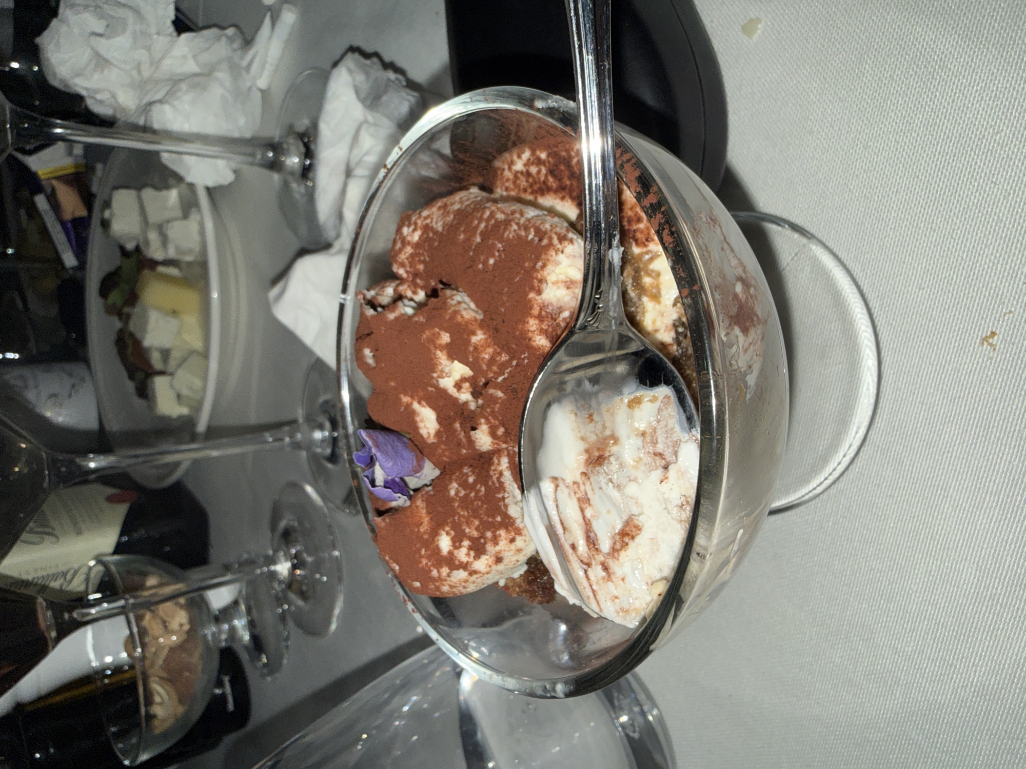 The image shows a dessert, likely tiramisu, in a clear glass bowl with a spoon in it. The dessert is dusted with cocoa powder and a small purple flower. The bowl sits on a table alongside wine glasses and other items, including what appears to be cheese cubes in a container. The background is light, suggesting a well-lit interior. The scene has the appearance of a meal on a table, possibly in a home or restaurant. There is no visible information to place the image in Kocherinovo, Bulgaria.