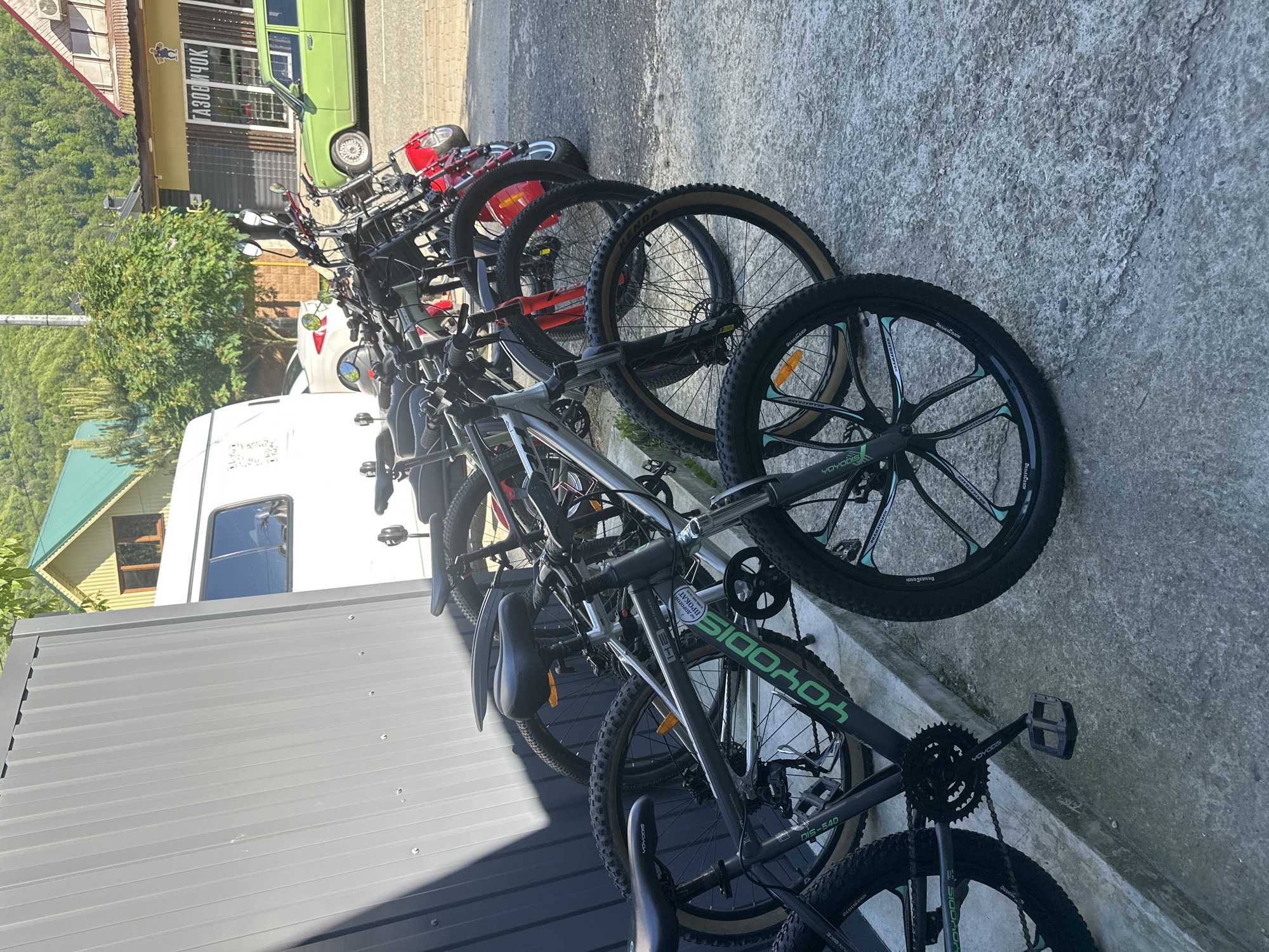 This outdoor photograph, set in Krasnaya Polyana, Russia, depicts a vibrant scene under clear, bright daylight. Numerous bicycles are neatly parked in a long line, suggesting a rental station or storage area. No individuals are visible.
The collection features a variety of mountain bikes. Several prominent black and green models bear the brand "YOYODIS" on their frames and feature distinctive five-spoke mag wheels. Other bikes have traditional spoked wheels, with "KENDA" clearly visible on some tire sidewalls and "DRAKCARSH" on others. A specific model, "DIS-540," is also noted on one frame. One bicycle sports a white sticker that reads "ЖИВОЙ РУЧОК" (ZHIVOY RUCHOK). Smaller, red-colored bicycles, likely intended for children, are also part of the lineup.
The bikes are parked on a textured concrete surface alongside a corrugated grey metal fence or wall. In the mid-ground, a vintage-style green car, resembling an SUV, is parked in front of a yellow-facade building. This building's storefront displays the Cyrillic text "ГАЗОВЩИК" (GAZOVSHCHIK) above a dark window. A white van or camper is partially visible behind some of the bikes, and beyond it, a house with a green roof can be seen. The background is dominated by lush green trees and distant mountains, characteristic of a scenic mountain resort location. The strong, crisp shadows cast by the bikes and other objects indicate a high sun and clear weather. The overall impression is one of readiness for outdoor recreation in a picturesque setting.