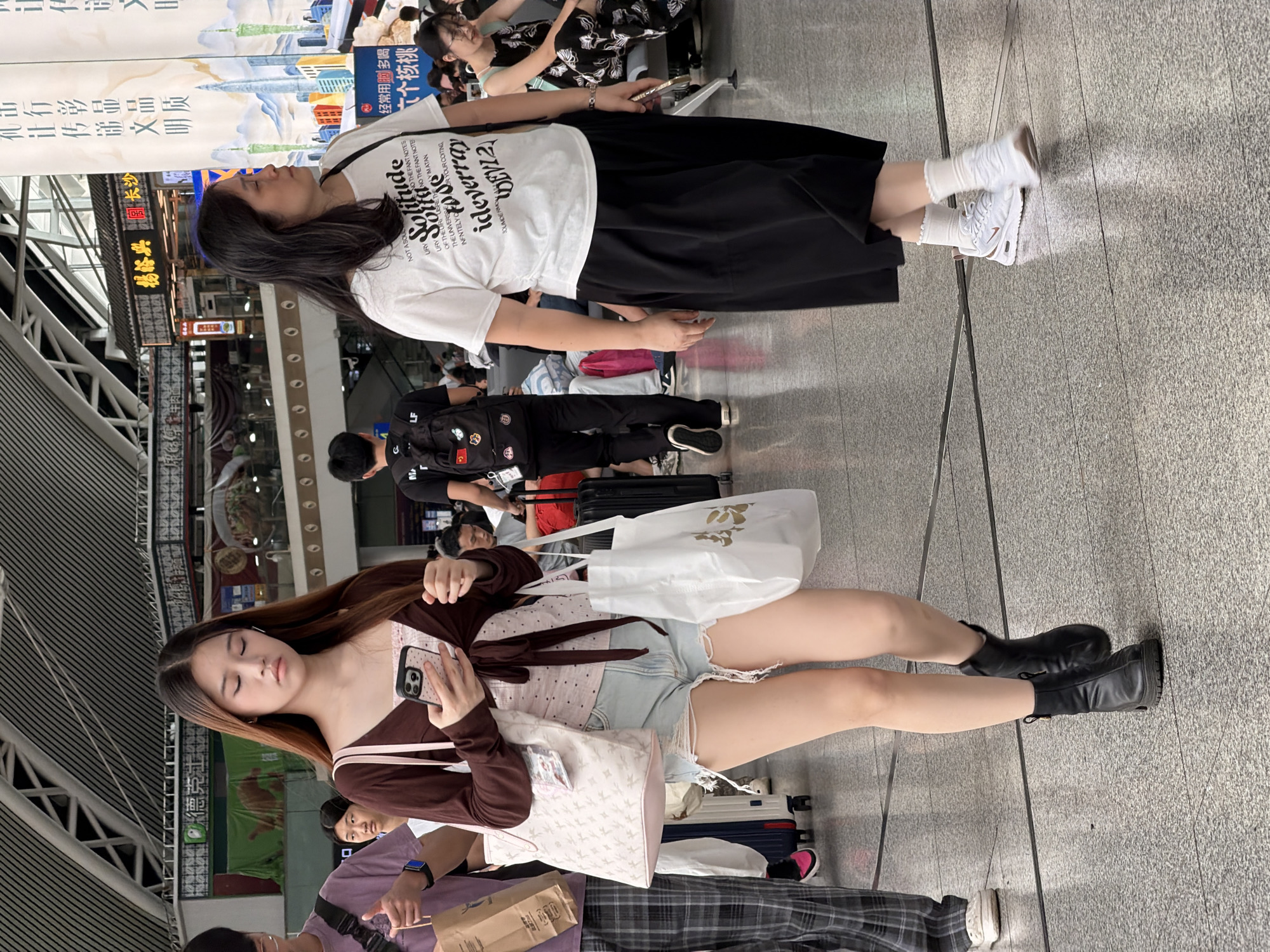 The image depicts a busy indoor environment, likely a train station or airport terminal in Panyu District, China, characterized by a polished, speckled floor and a high ceiling with visible structural elements. In the foreground, a young woman with long brown hair is walking while looking at her phone. She is wearing a brown open cardigan over a pink top, ripped denim shorts, and black ankle boots. She is carrying a large white patterned tote bag over her left shoulder and a white shopping bag in her right hand. To her right, an adult woman with long black hair is walking. She is wearing a white t-shirt with text and graphics and a flowing black skirt, paired with white sneakers and white socks. Other individuals, including a person with a backpack and someone in plaid pants, are visible in the background, suggesting a public transit hub. Banners with Chinese characters hang from the ceiling, further indicating the location.