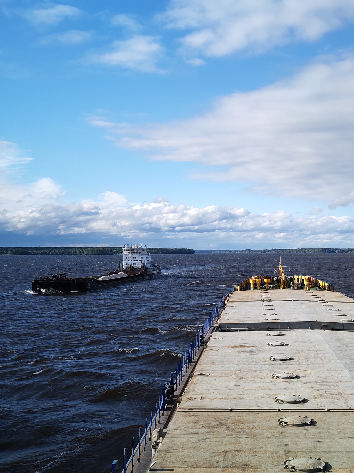 This is a vertical, medium shot photograph taken on a body of water, possibly the Sheksna River in Sheksna, Russia, on a partly cloudy day. In the foreground, the deck of a barge or similar vessel is visible, with a grey, weathered surface and numerous circular hatches or manholes. A blue railing lines the left edge of the deck. In the middle ground, a separate white and grey vessel, likely a tugboat pushing a barge, is traveling away from the viewer towards the left. This vessel has a multi-story superstructure and is laden with a light-colored material on its barge portion. The water is dark blue and choppy with whitecaps, suggesting wind. The background features a distant, tree-covered shoreline under a sky with white, fluffy clouds against a bright blue backdrop.