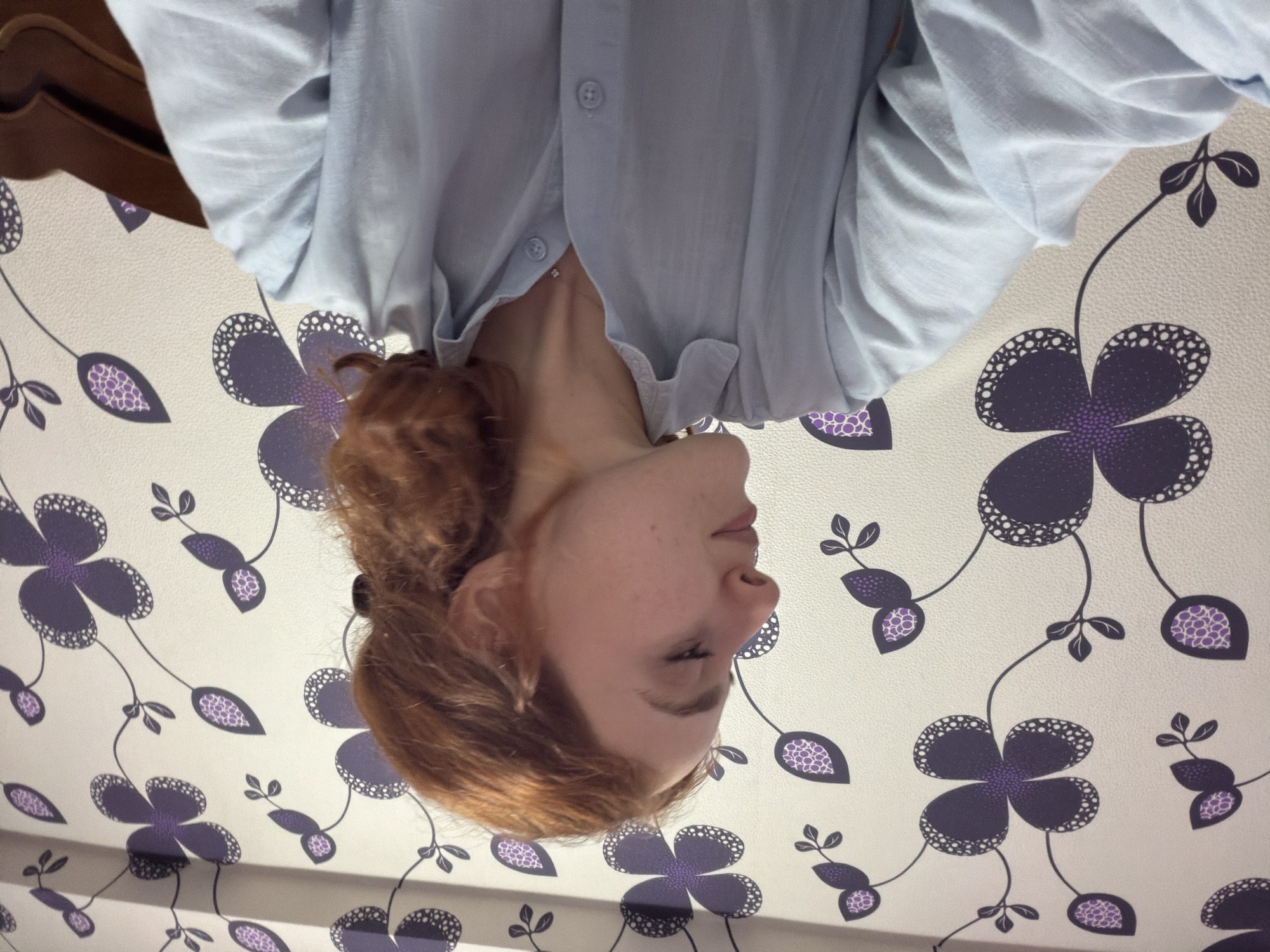 A woman with red hair is wearing a light blue, button-down shirt. She appears to be lying down, as indicated by the floral patterned wall behind her, which suggests the scene is indoors, possibly a bedroom. No information suggests this scene is in Tbilisi, Georgia.
