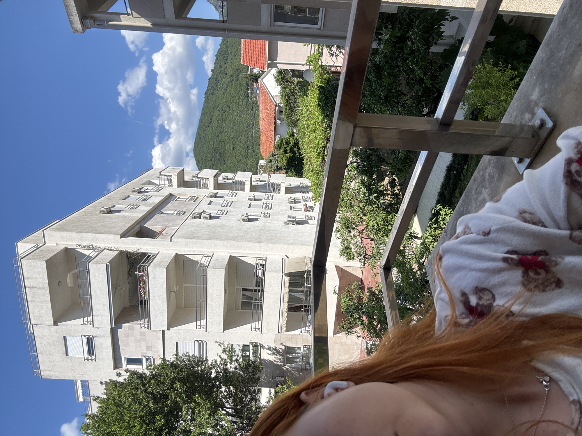 The scene is captured from a high vantage point, likely a balcony, offering a view of a residential area in Budva, Montenegro. A multi-story white building with numerous balconies occupies a significant portion of the view. In the background, a green, densely vegetated mountain rises, while a glimpse of a red-roofed building is visible amidst the greenery. A metal railing and part of a person with red hair in a white shirt with bear prints are in the foreground, suggesting the photograph was taken from their perspective. The sky is blue with scattered clouds, indicating a clear day.