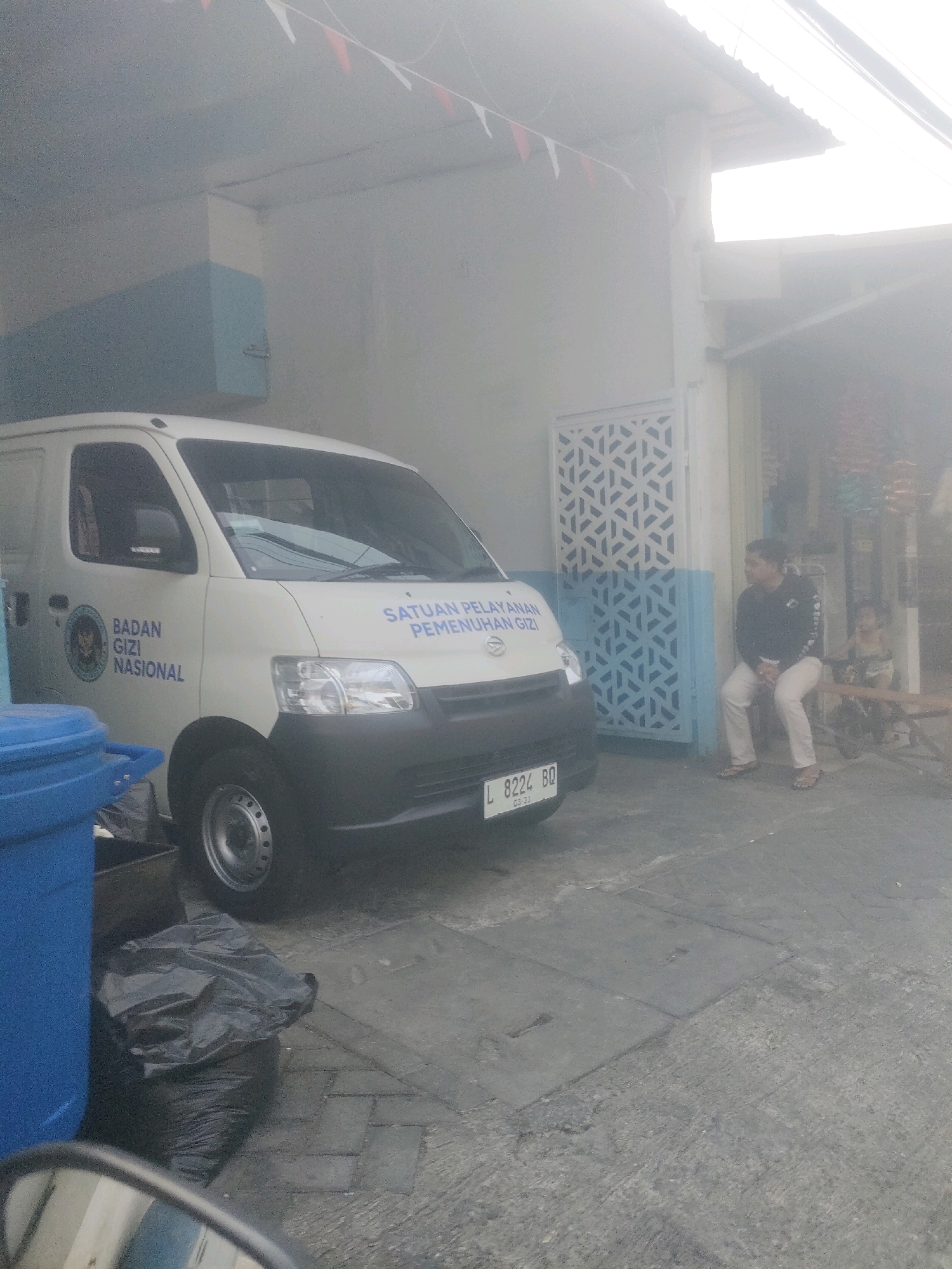 The image depicts a street scene in Surabaya, Indonesia, characterized by an overexposed, hazy atmosphere, likely due to bright sunlight or fog. In the foreground, a white van with blue lettering is parked. The lettering on the van reads "BADAN GIZI NASIONAL" and "SATUAN PELAYANAN PEMENUHAN GIZI". A blue trash bin is situated to the left of the van, with a black garbage bag placed on the ground beside it. A side mirror of what appears to be a vehicle is visible in the bottom left corner, reflecting a blurred image.
Further into the scene, a person is seated on a wooden bench outside a building with a distinctive patterned door. This person, wearing a dark jacket and light-colored pants, is looking to the right. Behind this person, a child is visible, also seated. The building itself has a white facade with light blue accents around the door. Decorative triangular flags are strung across the top of the building. The ground is paved with a mixture of concrete slabs and pavers. The license plate of the van is visible and reads "L 8224 BQ".