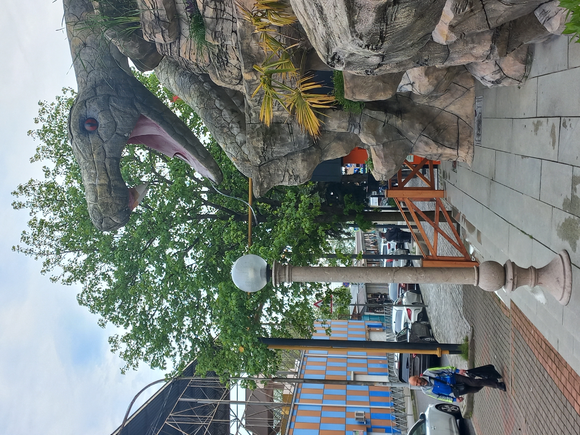 The image depicts a scene on a street in Sochi, Russia. Dominating the view is a large, decorative sculpture of a snake head emerging from artificial rocks and foliage. The sculpture is positioned above the street.
A street lamp stands near the sculpture. Behind the lamp and snake is a tree, partially obscuring the view of buildings. A building with a blue and orange checkered pattern is visible. Further down the street, cars are parked, and people are walking.
A man wearing a blue jacket is walking on the sidewalk near the street lamp. The sidewalk is paved with bricks. Wooden barriers are visible on the side of the street, bordering the sidewalk. Scaffolding is visible in the lower left corner of the image.