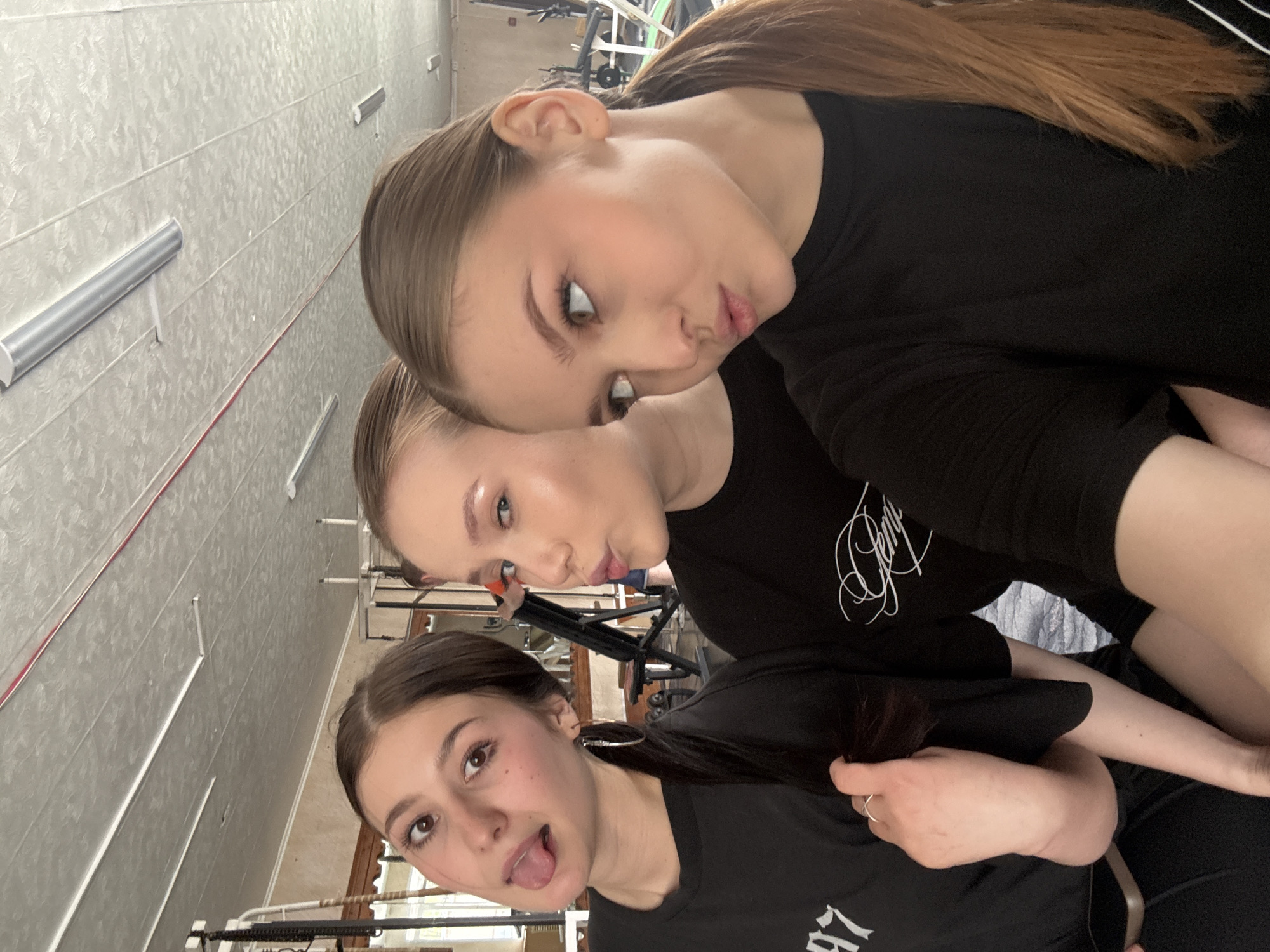 Three young women are posing close together for a selfie in an indoor space, likely a gym or dance studio, based on the equipment visible in the background. The topmost woman has light brown hair, styled back. She is making a duck-face expression. Below her, another woman is also posing with pursed lips. The woman in the foreground is sticking out her tongue playfully. All three women are wearing black shirts. The location is possibly in the city of Chaikovsky, Russia. The background includes workout equipment and a wall with metal fixtures.