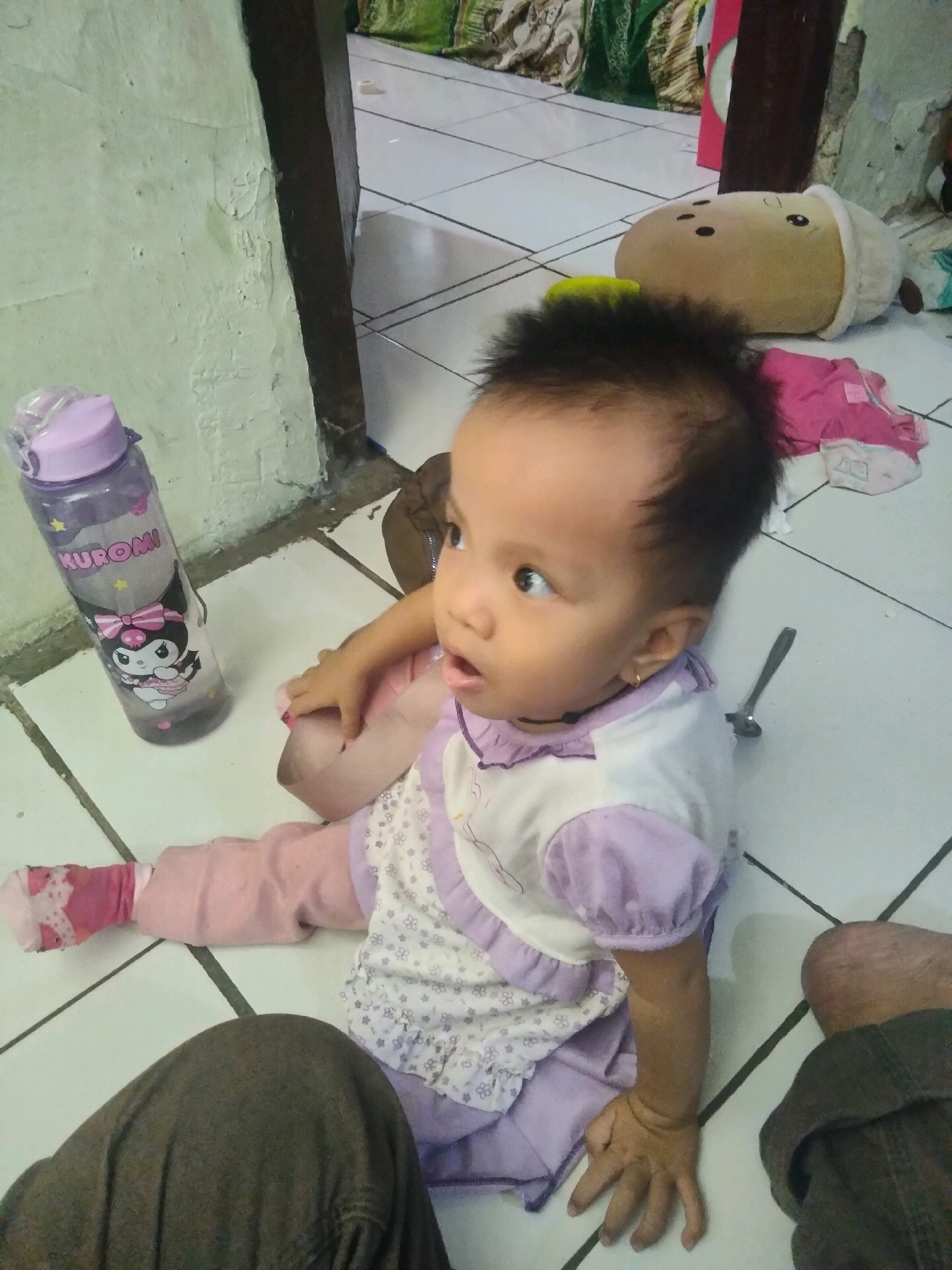 The image depicts a young child sitting on a tiled floor in what appears to be a room with white, cream, and green walls. To the left, a purple water bottle with a cartoon character and the name "KUROMI" is visible. The child is wearing a white shirt with purple trim and a purple skirt, with pink socks. A person's legs, clad in dark trousers, are in the lower part of the frame. A stuffed toy resembling a round, brown character with black dots for eyes and a white hat is on the floor behind the child. A small metal object, possibly a spoon or a similar utensil, lies on the floor near the child's right side. The overall environment suggests a domestic setting, potentially in Suka Sari, Indonesia, given the context provided.