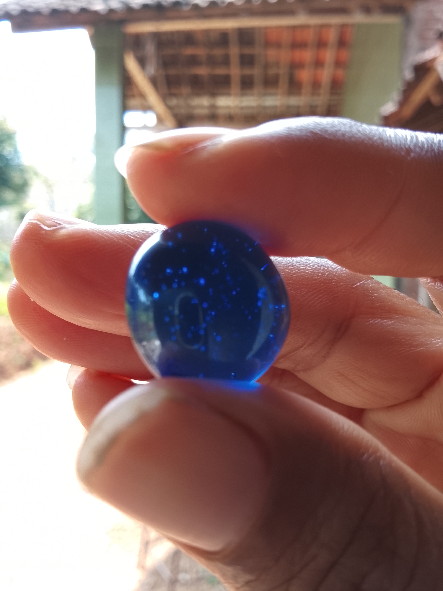 In Dawung, Indonesia, a close-up shot focuses on a person's hands holding a translucent, sapphire-blue marble. The marble is spherical and appears to contain small, reflective flecks scattered within its depths. The fingers and thumb are gently cupping the marble, with some fingernails visible and neatly trimmed. The background is softly blurred, revealing the indistinct outlines of a building with a tiled roof and wooden supports, suggesting an outdoor or semi-outdoor setting. The lighting suggests it is daytime, with bright light filtering in from the left, illuminating the marble and the hands. The overall impression is one of careful handling of a small, decorative object.