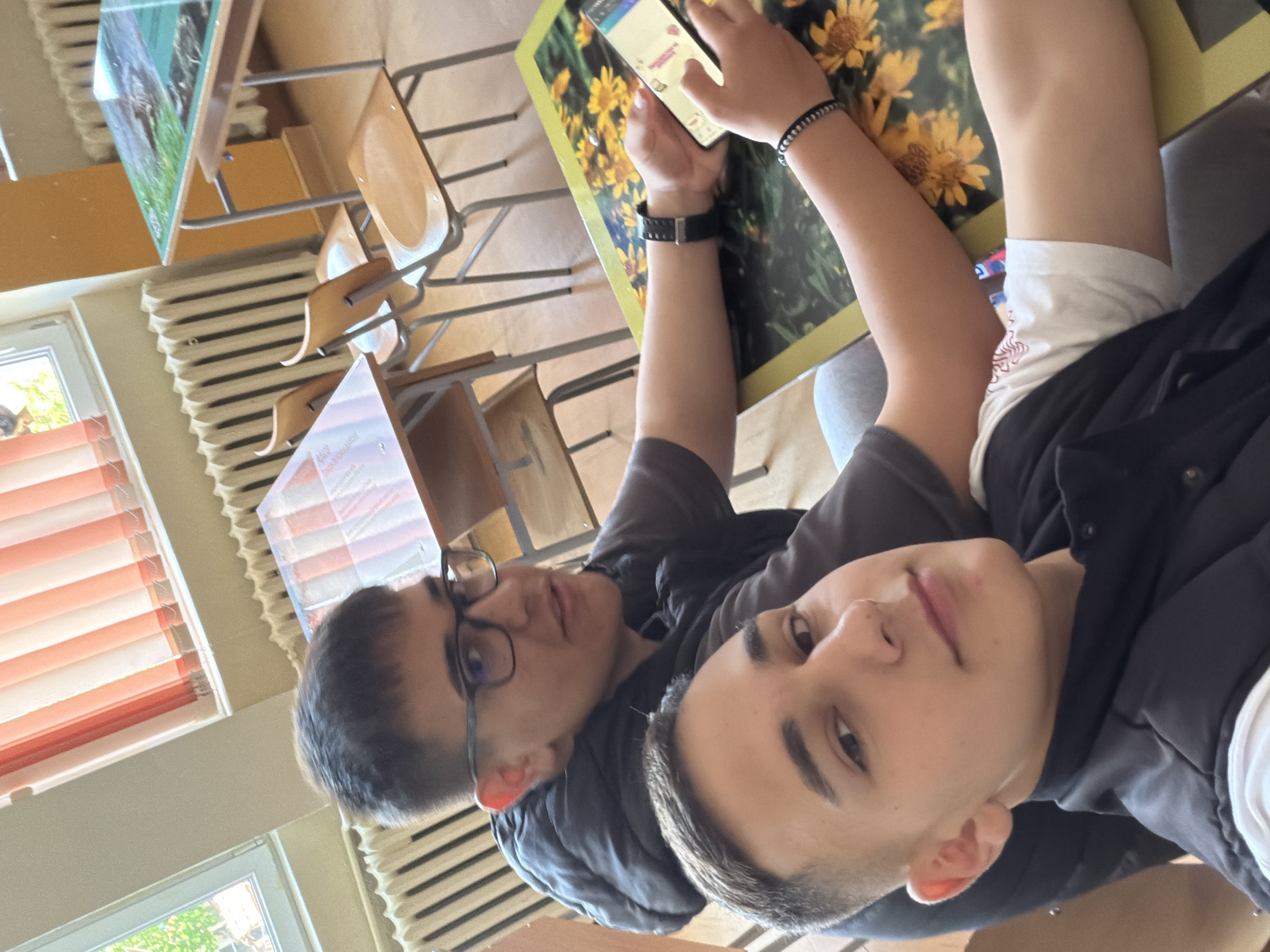 Two young males are posing indoors. One, wearing glasses, is looking at the camera; the other is looking upwards while holding a smartphone. They are situated among desks and chairs, with textbooks and printed materials visible on the desks. A picture of flowers is visible. The background indicates a classroom setting with orange blinds on the window and visible radiators. The location is likely a school in Panagyurishte, Bulgaria.