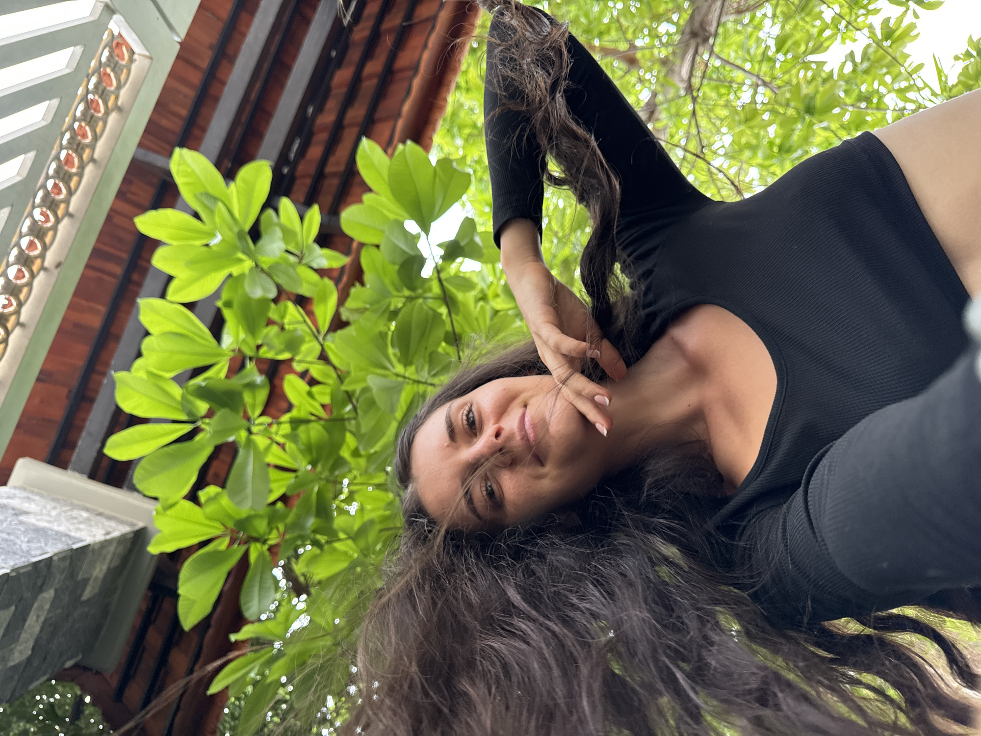 The image presents a low-angle portrait of a woman, likely taken outdoors in Mũi Né, Vietnam. The main subject is a woman with long, dark, wavy hair, which frames her face and flows downwards towards the bottom of the frame. She is wearing a form-fitting black ribbed top or bodysuit. Her left hand is subtly raised, with her fingers touching her hair near her cheek, and she looks directly at the camera with a gentle, pleasant smile.
The background is a mix of natural and architectural elements. Lush, bright green foliage from a tree or shrub occupies a significant portion of the upper and left side of the frame, indicating a sunny, outdoor setting. Behind the greenery, elements of a building are visible: an orange-brown tiled roof with dark support beams, typical of local construction. To the far left, a light-colored architectural detail with a patterned design, possibly a decorative frieze or railing, is partially seen, along with a grey, stone-like pillar in the bottom-left corner. The lighting suggests bright daylight, and the overall mood conveyed by the woman's expression is one of ease and contentment. There is no visible text in the image.