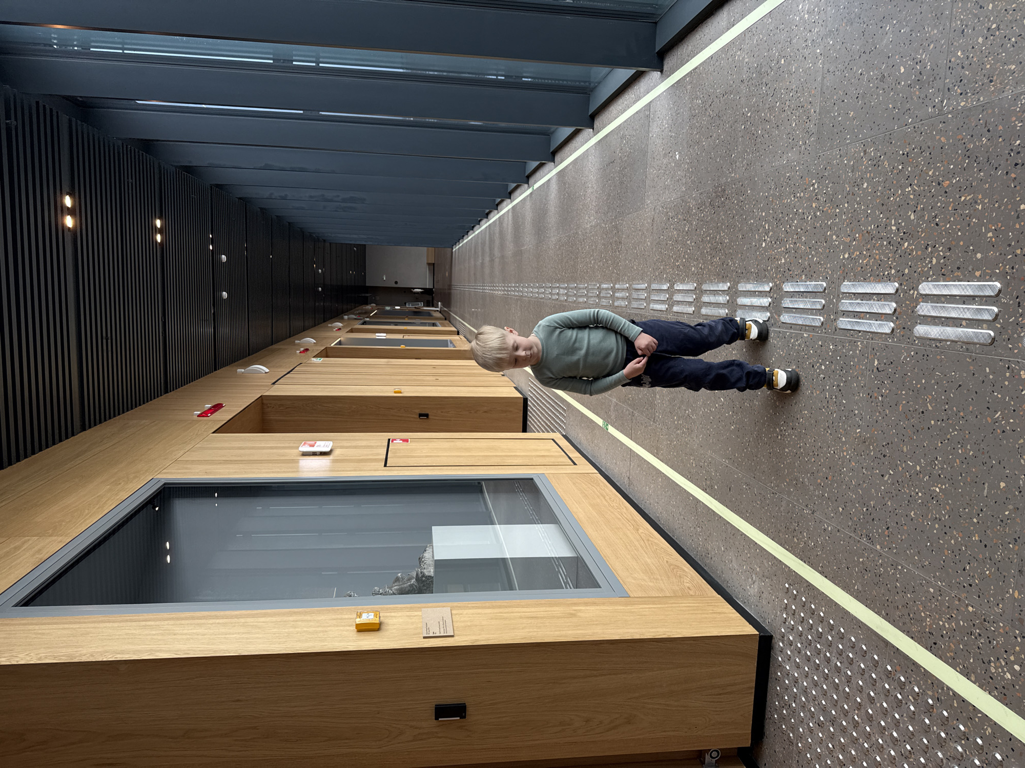 A full eye-level shot depicts a young child standing in a long hallway. The hallway features a stone floor with tactile paving, a yellow stripe indicating a pathway, and display cases recessed into the floor. The child is wearing dark pants, a light green long-sleeved shirt, and dark shoes with beige soles. The display cases have glass tops and appear to contain exhibits. The walls on the left are dark and vertical with inset lighting, while the walls on the right are the same stone material as the floor. Skylights run the length of the ceiling. The location is the Perm Museum of Contemporary Art, located in Perm, Russia.