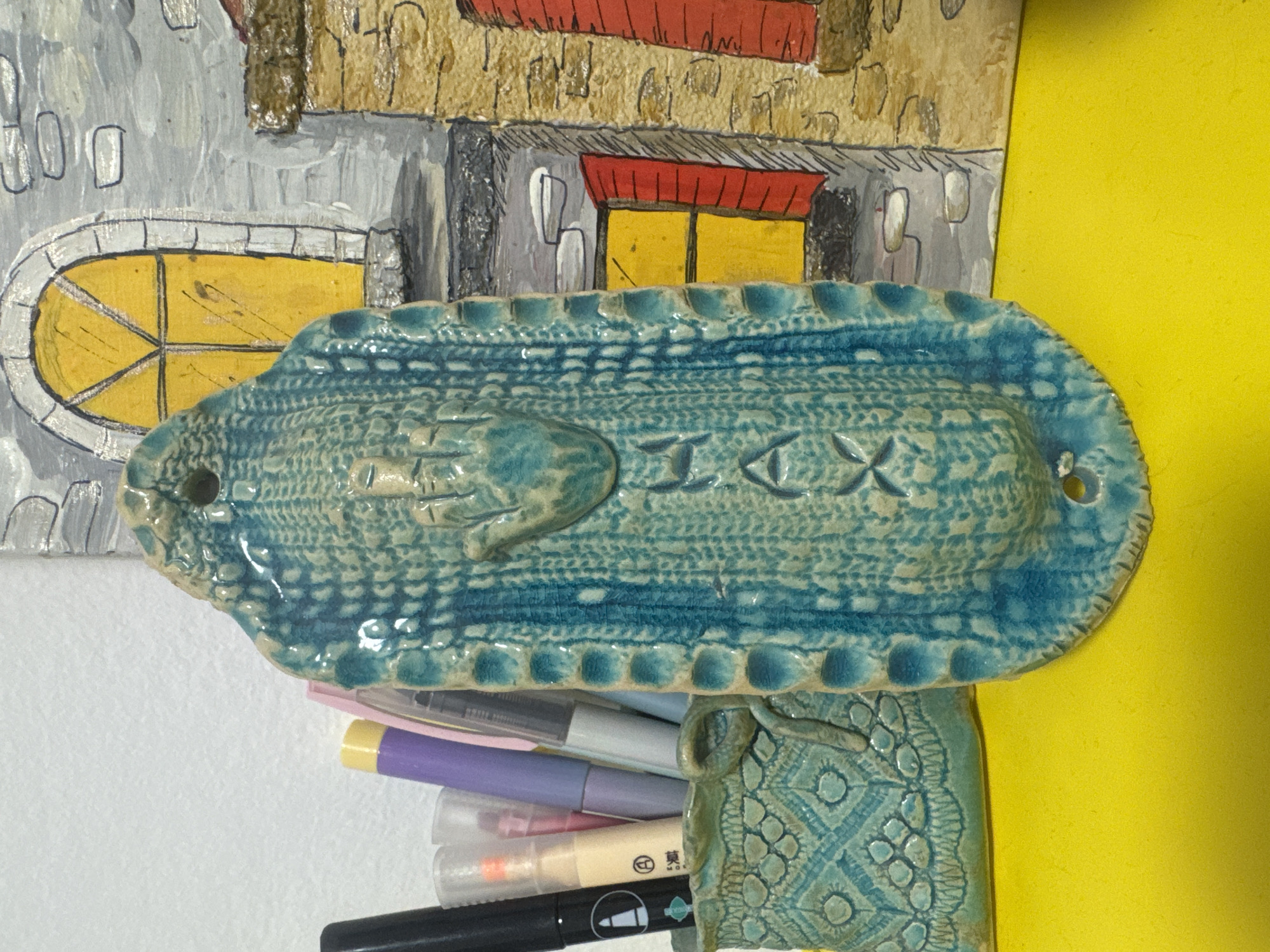 The image shows a close-up of a textured, blue ceramic decorative object, possibly a wall hanging or a planter, with a raised hand motif and some incised geometric patterns. It is positioned in front of a painted backdrop that resembles buildings with windows and doors. To the left of the ceramic piece, a basket-like ceramic container holds various pens and markers, including black, beige, purple, and gray ones. The background and foreground are brightly lit, with a yellow surface visible at the bottom. The overall scene appears to be an indoor display, possibly in an art studio or shop, located in Ramat Gan, Israel. There are no people visible in the image.