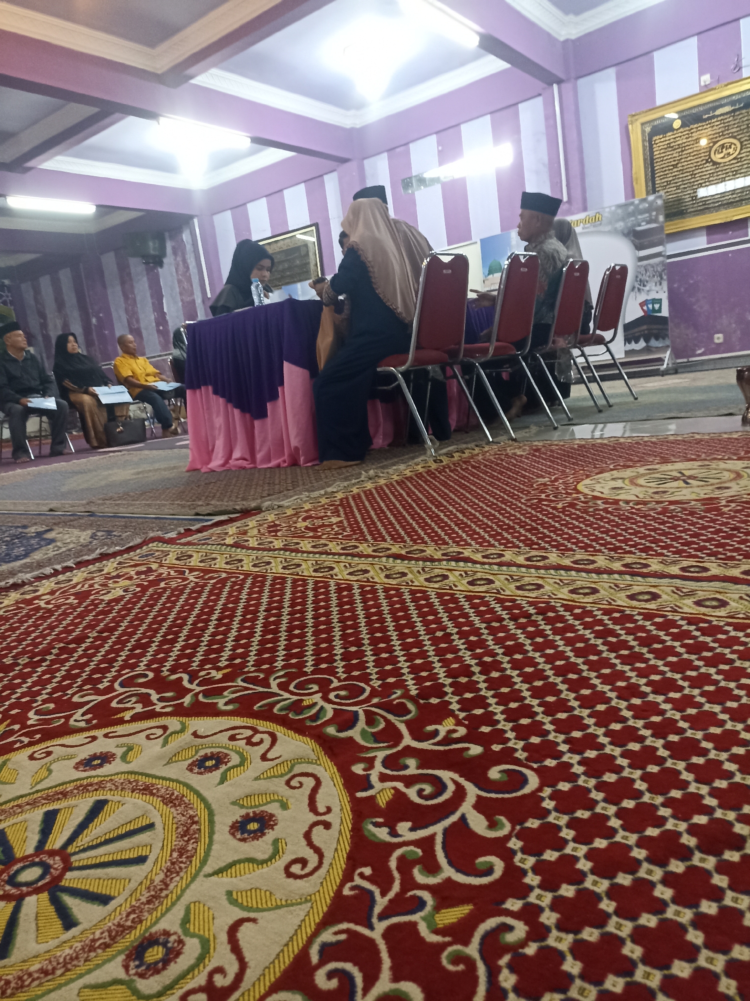 The image depicts an indoor gathering, likely a ceremony or meeting, taking place on a large, intricately patterned red carpet. The carpet features a central circular medallion with floral and geometric motifs in cream, yellow, blue, and green, bordered by a repeating pattern of small red and white quatrefoils. In the background, a group of people are seated or standing around a table draped with purple and pink fabric. Several individuals are wearing religious attire, including hijabs and skullcaps. A framed Islamic calligraphy artwork hangs on a purple and white striped wall. The overall atmosphere suggests a formal or community event in a decorated indoor space, possibly a mosque or community hall in Tompokersari, Indonesia.