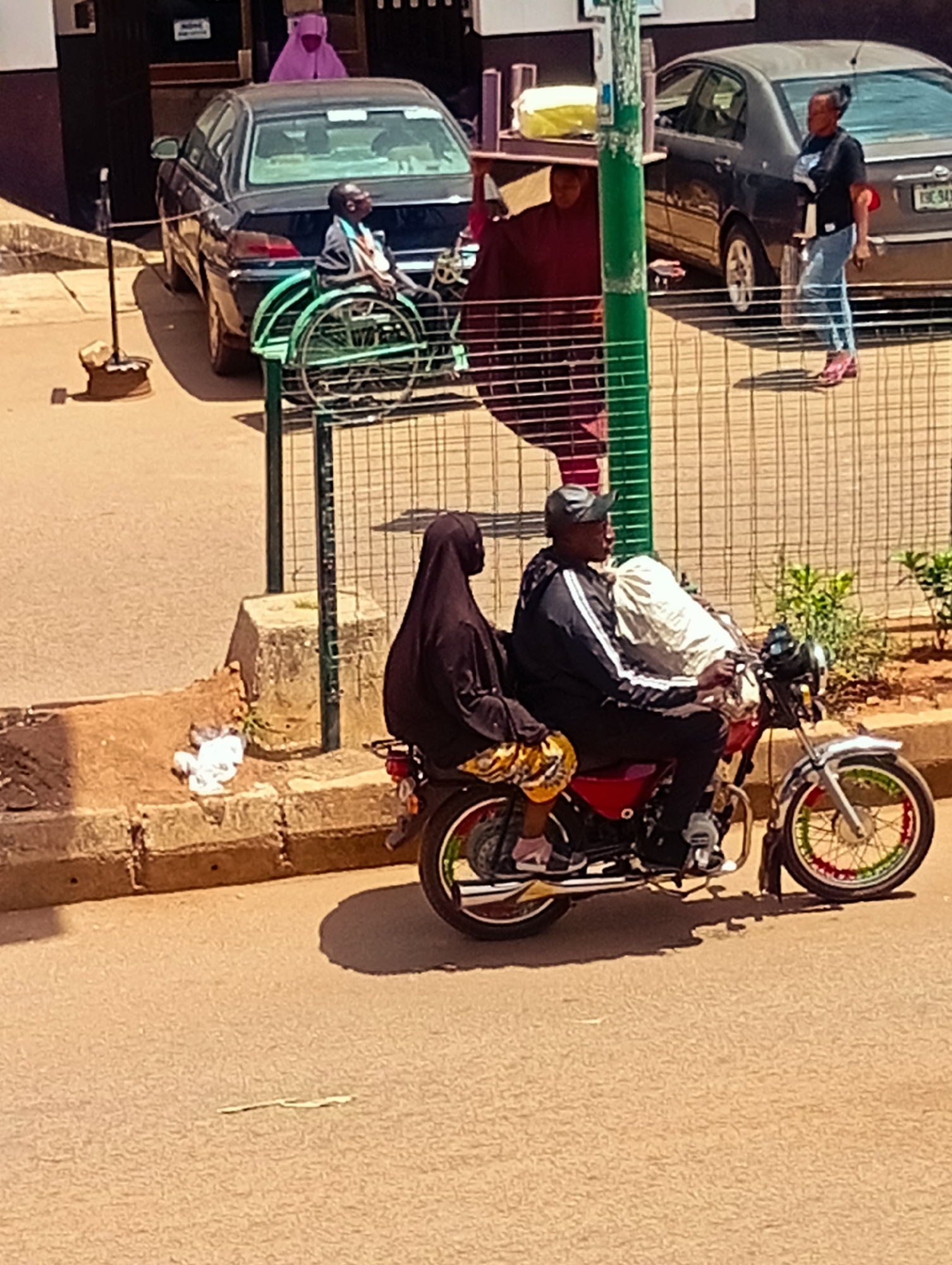 In Bukuru, Nigeria, two individuals are on a red motorcycle. The person in the back, wearing a black hijab and yellow and gold patterned skirt, is seated. The person in front, a male wearing a black baseball cap and a black jacket with white stripes, is driving. The motorcycle is parked on a paved street next to a dirt embankment. In the background, a dark sedan is parked, with a person in a wheelchair positioned next to it, behind a metal fence. Further back, another person in a purple garment stands near a building. The scene is outdoors, under daylight conditions.