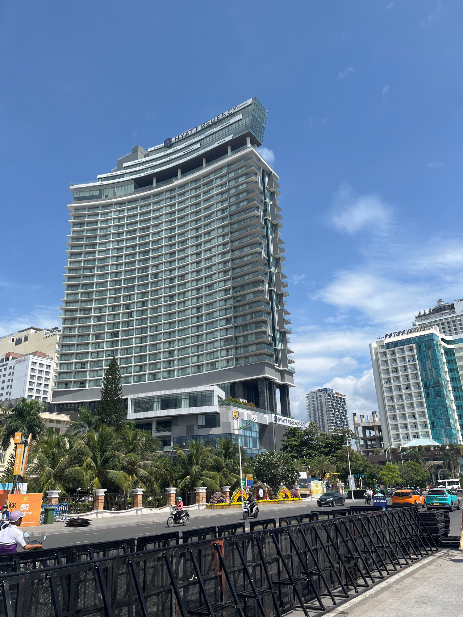 The image captures a street scene in Nha Trang, Vietnam, with a prominent view of the Hyatt Regency hotel. The hotel, a large, modern building with a distinctive curved facade and a glass-enclosed top section, dominates the skyline. In front of the hotel, a row of palm trees lines the sidewalk. Across the street, several vehicles, including motorcycles and cars, are visible. The street is lined with temporary black barricades. Signs in Vietnamese are present, indicating a commercial area. The sky is blue with scattered white clouds, suggesting a sunny day. The scene depicts a typical urban environment in a coastal city.