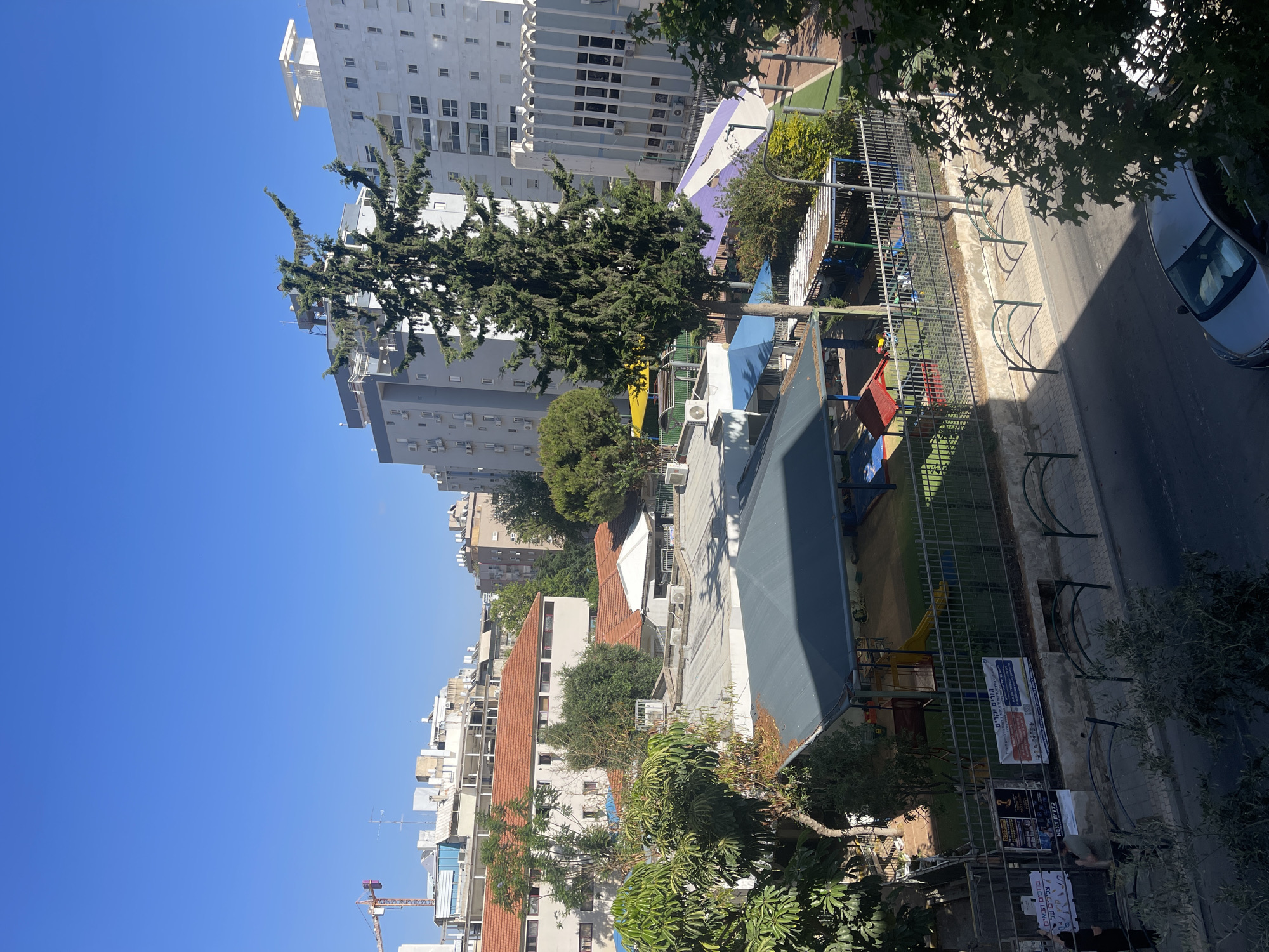 The image captures a view of a city scene in Netanya, Israel, with a clear blue sky. In the foreground, a street with a silver car is visible. A metal fence runs along the edge of the sidewalk, separating it from a grassy area and a playground. The playground features a covered sandbox, a slide, and colorful play structures. Beyond the playground, a tall evergreen tree stands prominently, with a backdrop of apartment buildings. The buildings vary in architectural style, some with orange-tiled roofs and others with modern facades. In the distance, a construction crane is visible, suggesting ongoing development in the area. Several trees and green spaces are interspersed throughout the urban landscape. There are a few people visible in the image, some near the fence and others further in the background. Posters are affixed to the wall behind the fence.