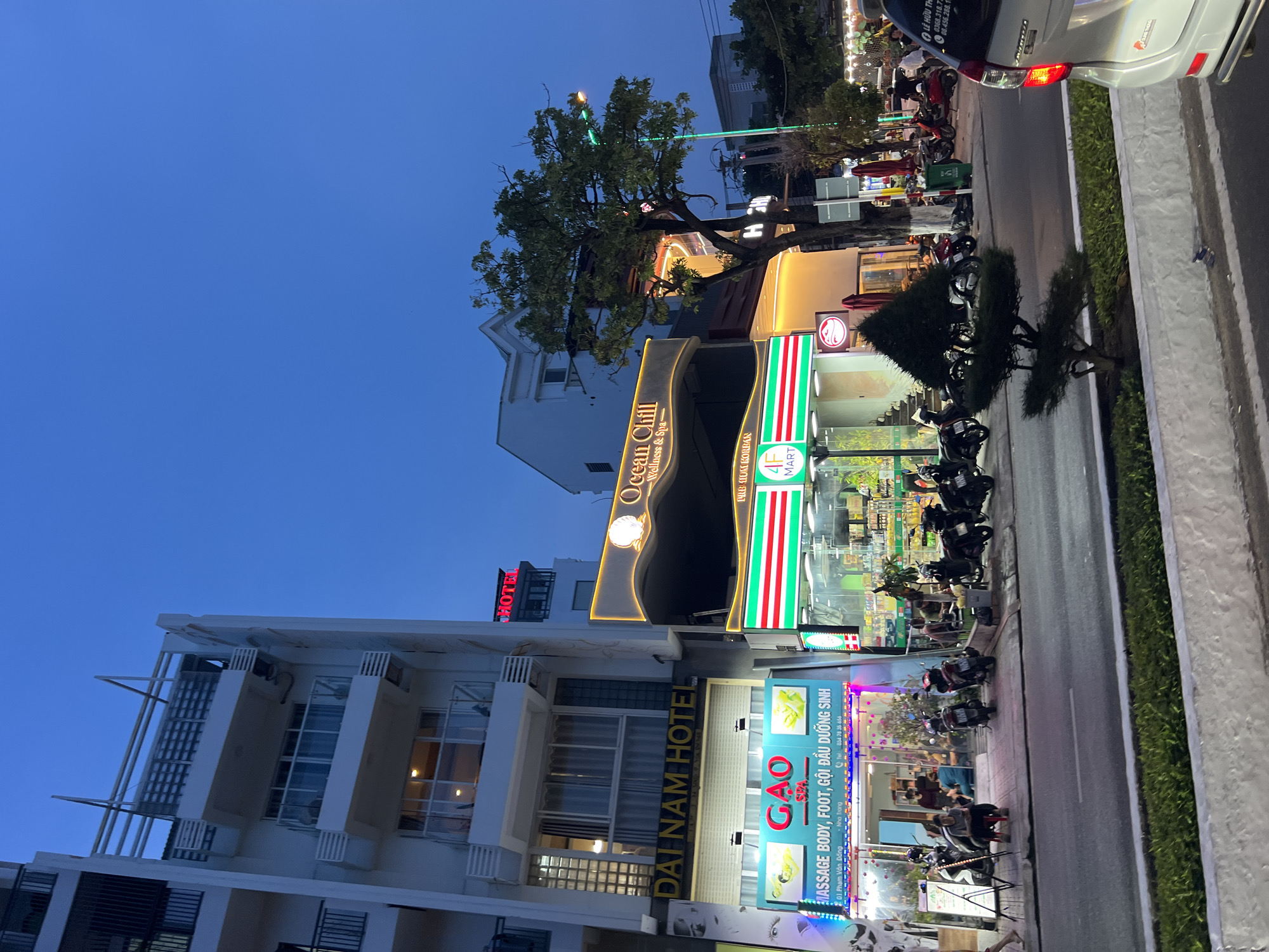 This image shows a street in Bac Nha Trang Ward, Vietnam, at dusk. The street is lined with buildings, including the Dai Nam Hotel, Ocean Chill Wellness & Spa, and a 4F MART. Motorbikes are parked along the side of the road, and a white car is partially visible at the top right of the frame. Streetlights and illuminated signs are lit, and the sky is a deep blue.