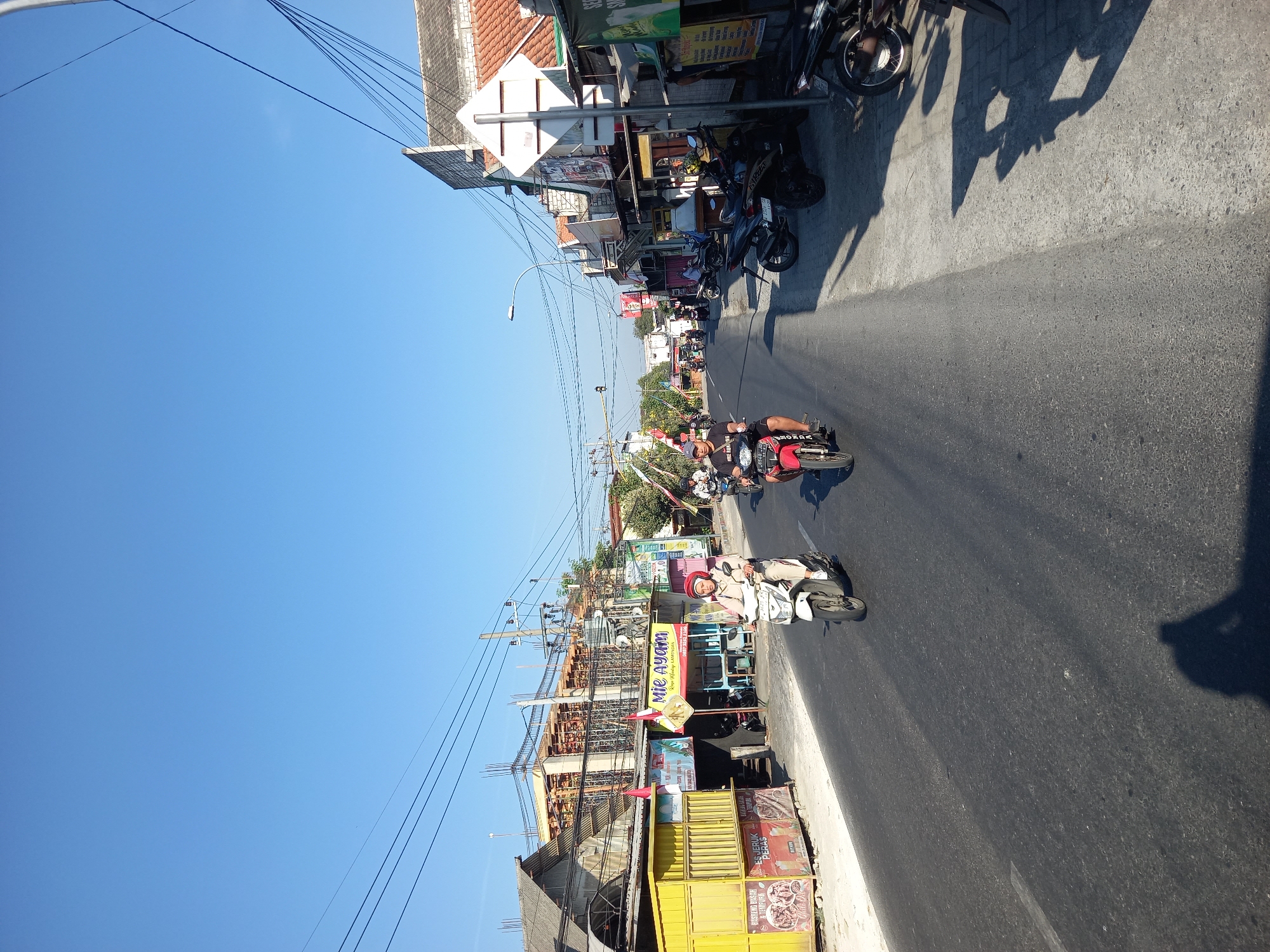 The image depicts a street scene in Bungah, Indonesia, likely captured during daylight hours. The foreground shows an asphalt road with a white lane marking. Two individuals on motorcycles are traveling down the road. The person on the left is riding a white scooter and is wearing a light-colored headscarf and clothing. The person on the right is riding a red and black motorcycle and is wearing a black shirt and shorts. The background reveals a commercial street lined with buildings. On the left side of the road, there are yellow structures that appear to be food stalls or shops, with signs advertising "Mie Ayam" (chicken noodles) and "Es Jeruk Peras" (pressed orange juice). Further down the street, more buildings with awnings and signs are visible, suggesting a busy marketplace or commercial district. Overhead, numerous electrical wires stretch across the sky, supported by utility poles. The sky is clear and blue, indicating good weather. The overall scene conveys a typical urban environment in Indonesia, with active transportation and commercial activity.