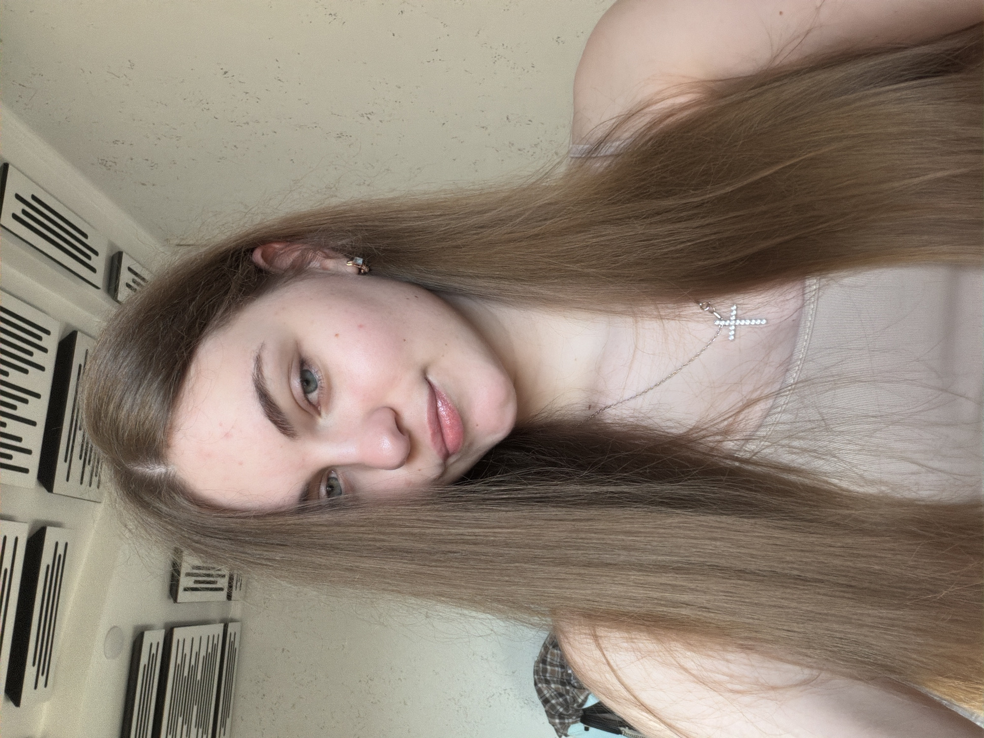This is a portrait of a young woman with long, light brown hair. She is wearing a necklace with a cross pendant and a simple tank top. Her expression is neutral and she is looking towards the camera. Her hair is the dominant feature in the image, falling around her face and shoulders. In the background, there are decorative wall panels with a black and white striped pattern. The lighting suggests it is daytime, and the overall setting appears to be indoors. The image has a slightly grainy quality. There is no visible text in the image.