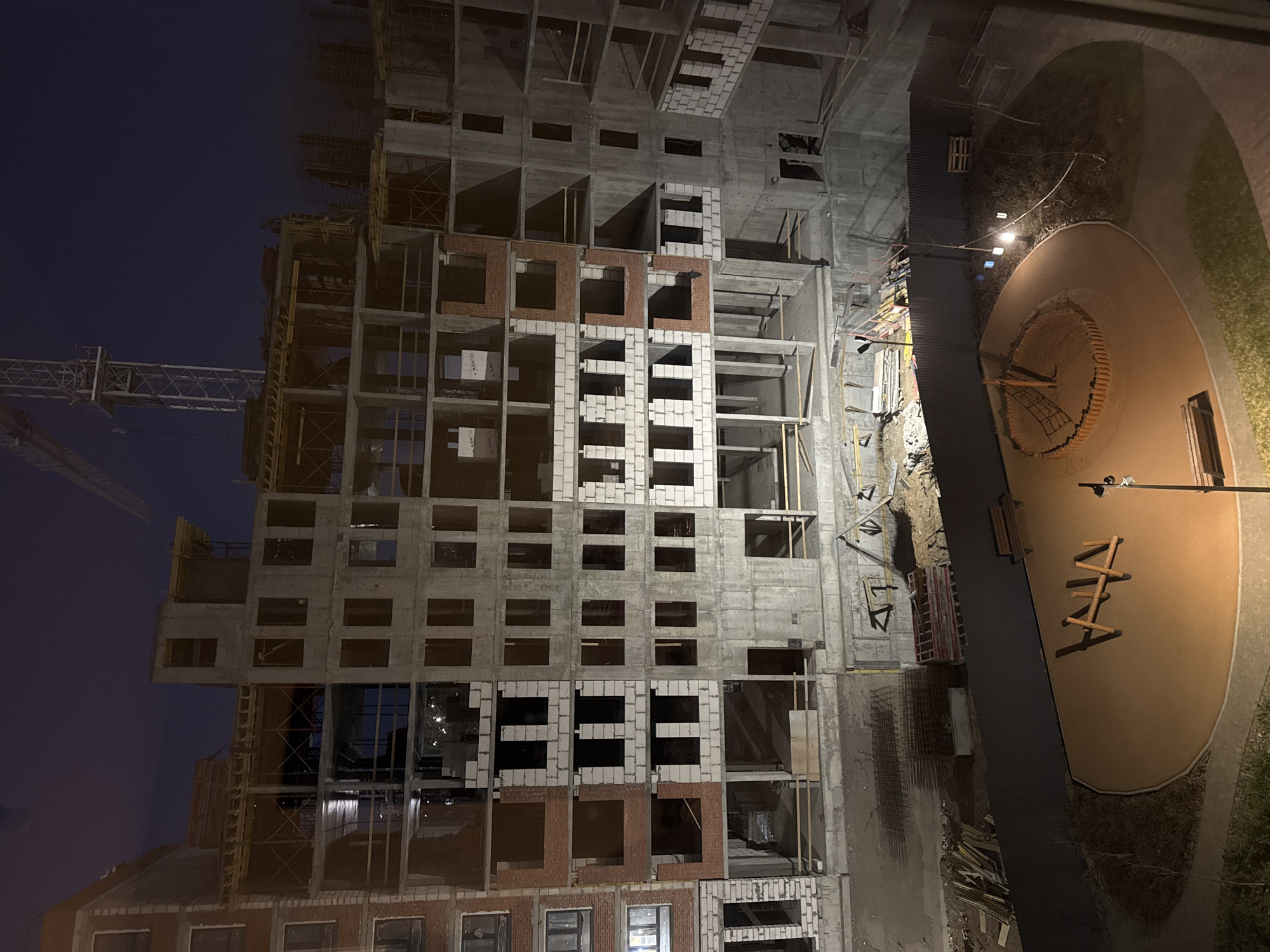 An aerial view of an uncompleted apartment building in Izhevsk, Russia, taken at night. The building, constructed of concrete and brick, is framed on the left by a tower crane under a dark blue sky. Adjacent to the construction site is a fenced playground.