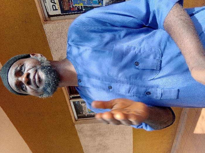 A man wearing a black skull cap and a light blue button-up shirt faces the camera with an outstretched hand. He has a graying beard. The setting appears to be outdoors with a yellow wall behind him, possibly the side of a building. A window with what seems like posters is visible.