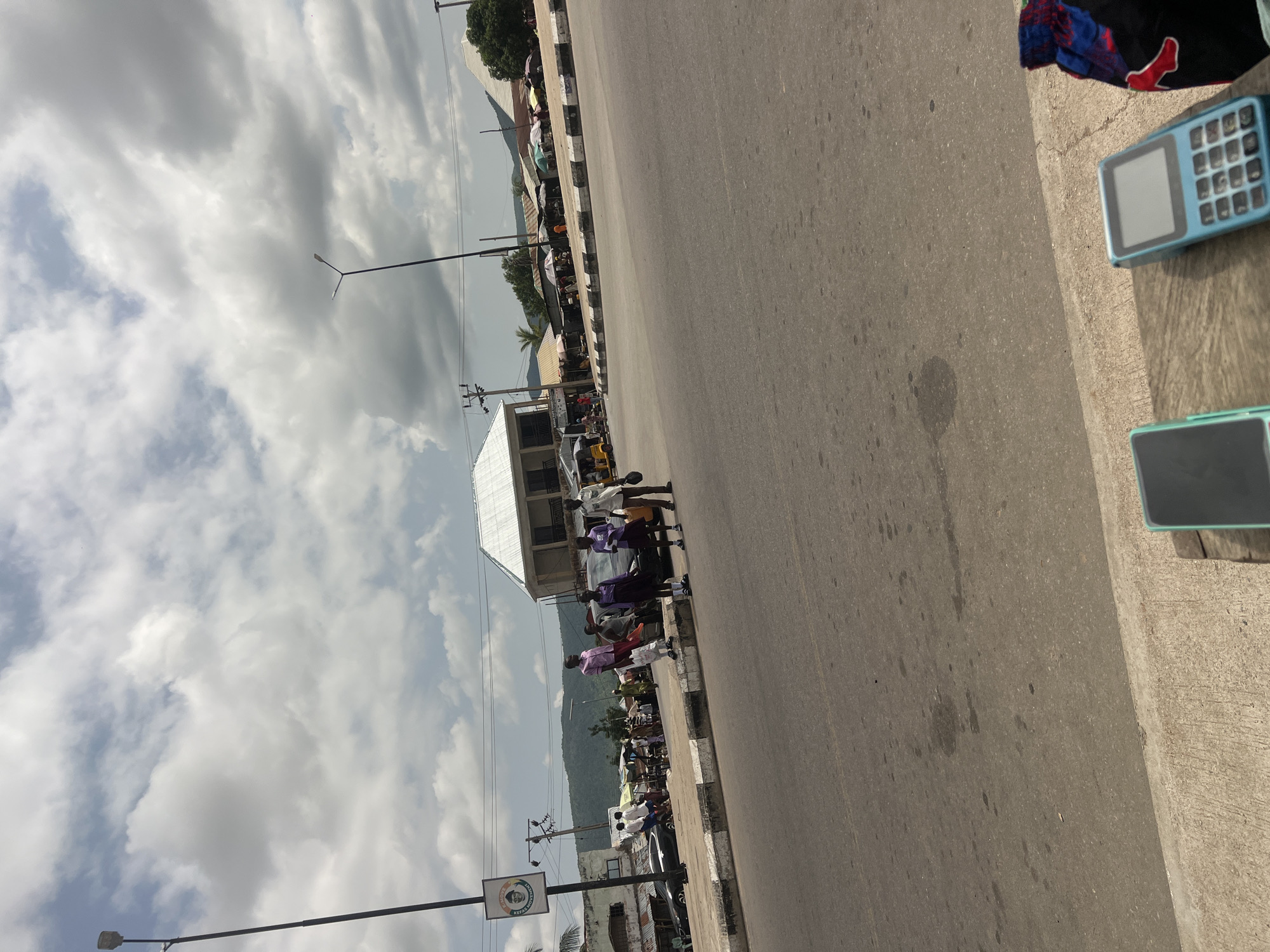 The media file captures a street scene in Howayi, Nigeria, likely during daytime with overcast skies. The perspective is low-angle, suggesting it was taken from ground level or slightly above.
The primary subjects are several people standing or walking along the side of a paved road. Many appear to be in casual attire, with some wearing shirts and shorts or trousers. In the foreground, to the right, are two POS (Point of Sale) terminals, one light blue and the other teal, resting on a concrete surface. A pile of dark-colored clothing with red accents is also visible near the POS terminals.
The setting is an urban or semi-urban area with a mix of structures. On the left side of the road, there are several stalls or small shops with awnings, and beyond them, a larger building with a white, sloped roof. Further back, a dense line of trees and a mountain range are visible under the cloudy sky. A streetlamp is present on the left side of the road, and electrical poles with wires are also visible. The road itself is asphalt, showing signs of wear and some dark stains.
The activity appears to be everyday street life, possibly a market area with vendors and pedestrians. The mood seems neutral, with no strong indicators of specific emotions.
There is some visible text on a circular sign on a pole near the lower left of the image. The text is small and difficult to read clearly, but it appears to contain some lettering and possibly a person's face in the center.
The presence of POS terminals might suggest commercial activity. The overall impression is of a busy, possibly commercial, street in a Nigerian town, with people going about their daily activities.