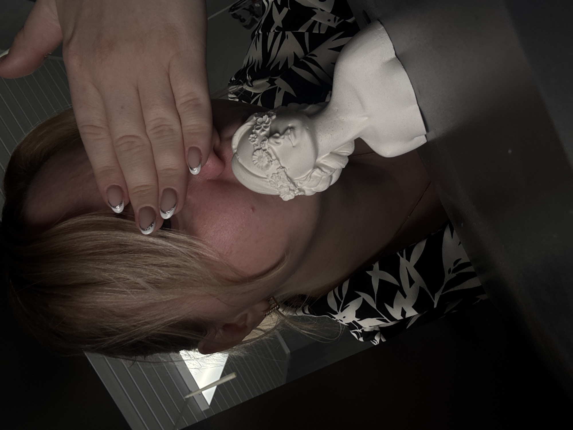 The image shows a person with blonde hair and fair skin covering part of their face with their hand. They have manicured nails with a white tip and silver glitter accent. The person is wearing a black and white floral patterned top. A white plaster bust of a female figure is positioned near their face. The background includes a grey wall and a glimpse of what appears to be blinds or a similar window covering. No information is available to confirm the location as Chaikovsky, Russia, or any other specific city.