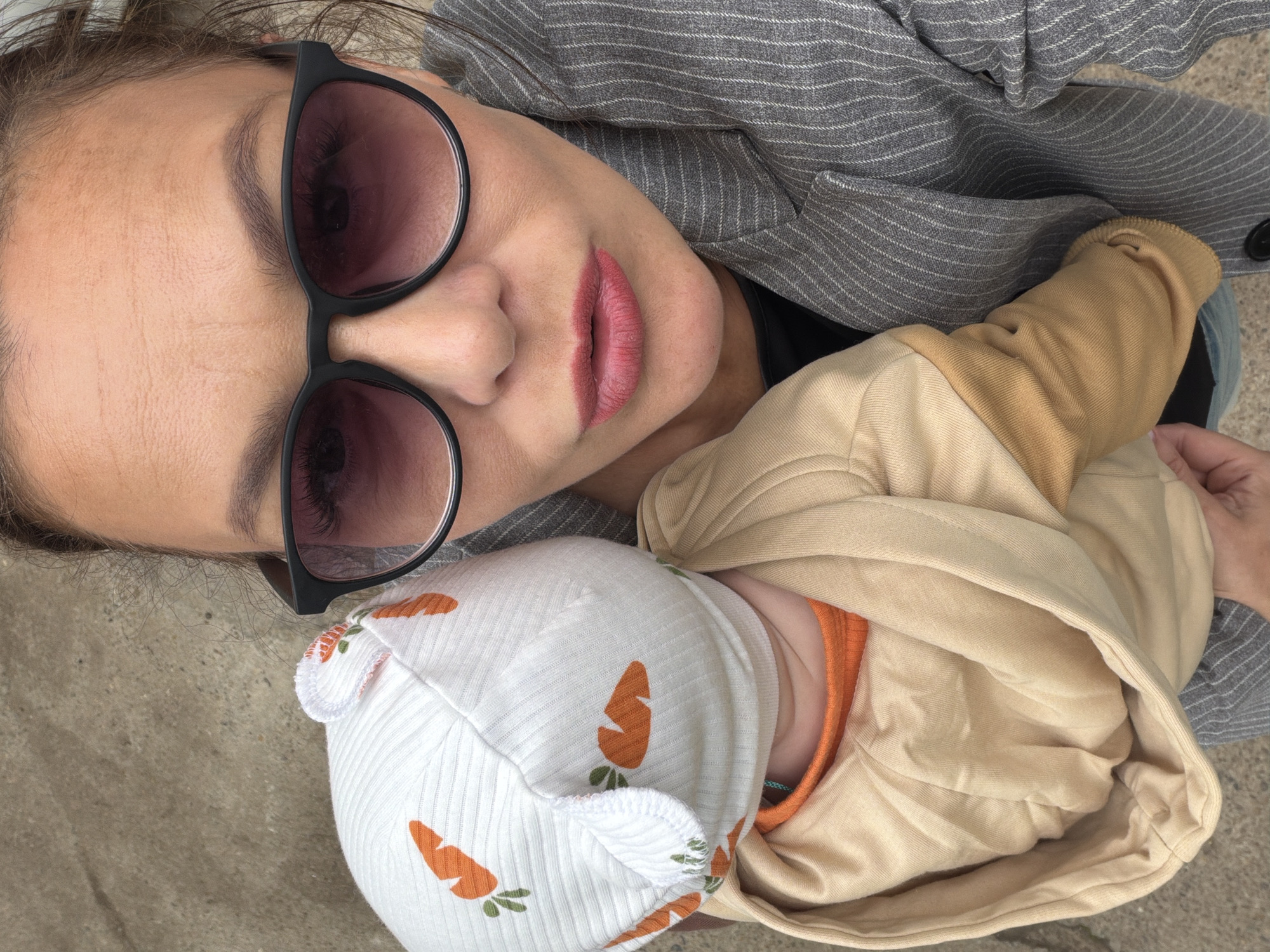 An overhead, close-up selfie shows a woman and a baby. The woman, wearing sunglasses and a pinstripe jacket, holds the baby, who is wearing a white hat with orange carrots and a tan hooded sweater over an orange shirt. The background appears to be an outdoor concrete surface. The image does not contain any specific landmarks or indicators to determine its location within Chaikovsky, Russia.