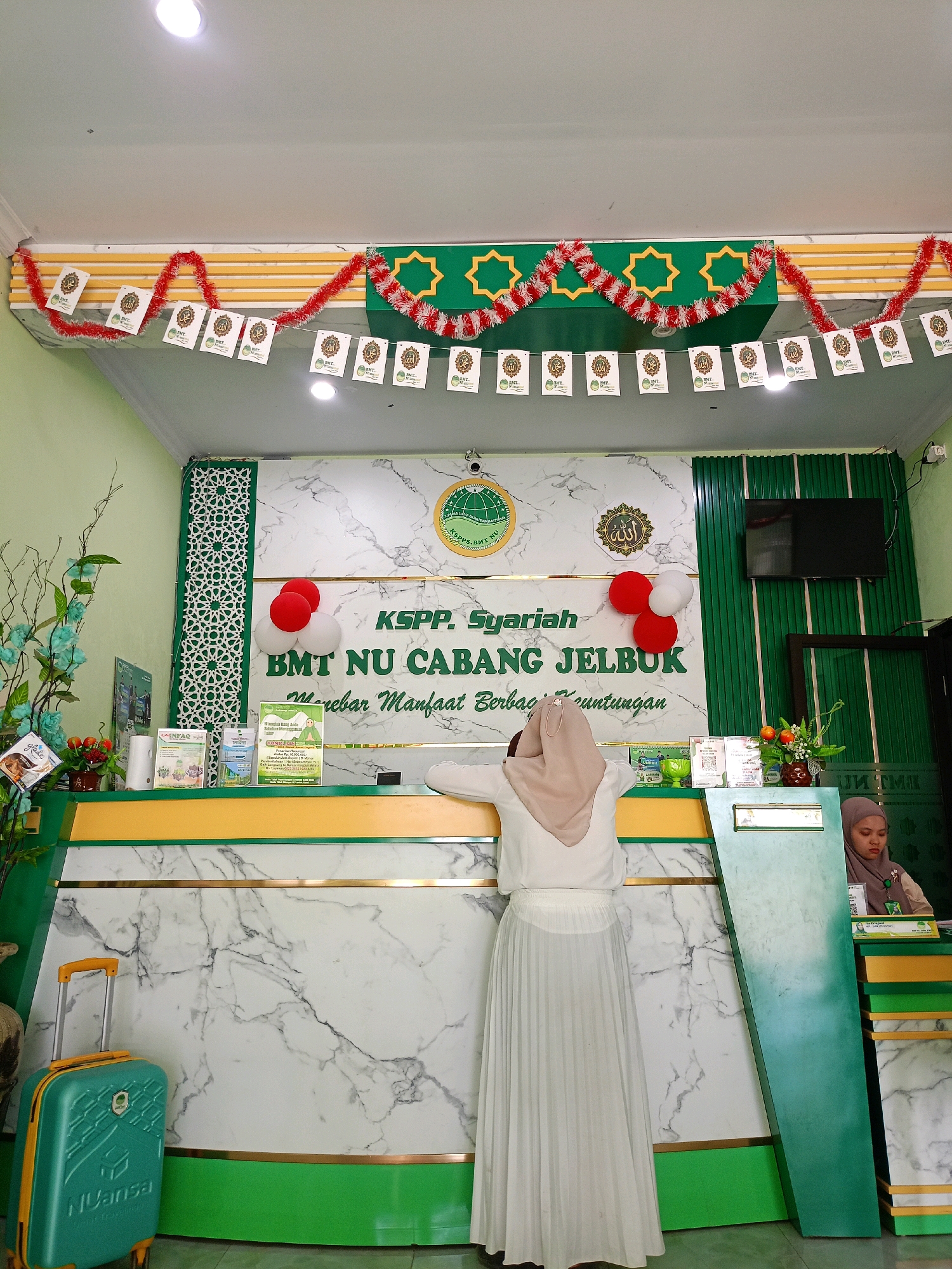 The image depicts the interior of a BMT NU Cabang Jelbuk office, likely a financial institution, in Suko, Jember, Indonesia. The scene is brightly lit, with a green and white color scheme dominating the decor. A counter with a marble-patterned front serves as the focal point, behind which two women are visible. One woman, wearing a hijab and a white outfit, stands facing the counter. The other woman, also wearing a hijab and a muted brown uniform, is seated behind the counter, appearing to be an employee.
Decorative elements include red and white tinsel garlands strung across the top of the counter, along with a banner featuring Islamic calligraphy and the institution's logo. Several brochures and pamphlets are displayed on the counter, suggesting information dissemination. A green suitcase with an orange handle stands to the left of the counter. The environment appears to be an indoor office space, possibly a customer service area.
