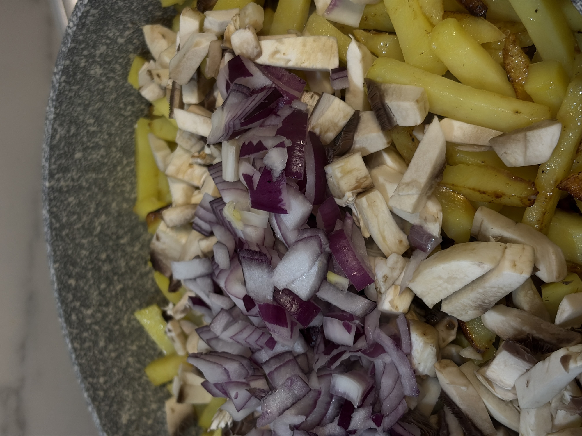 A close-up shot from above shows a gray-speckled pan filled with raw ingredients for cooking. Dominating the central area are chopped red onions. Adjacent to the onions are sliced mushrooms, predominantly white and gray in color. Along one side of the pan are cooked yellow potatoes, some with browned edges. The pan is sitting on a white countertop. The image contains no people or action, only a collection of food items preparing for cooking. There is no basis to conclude that it is taken in the city of Krasnodar, Russia, as that information is not available in the given context.