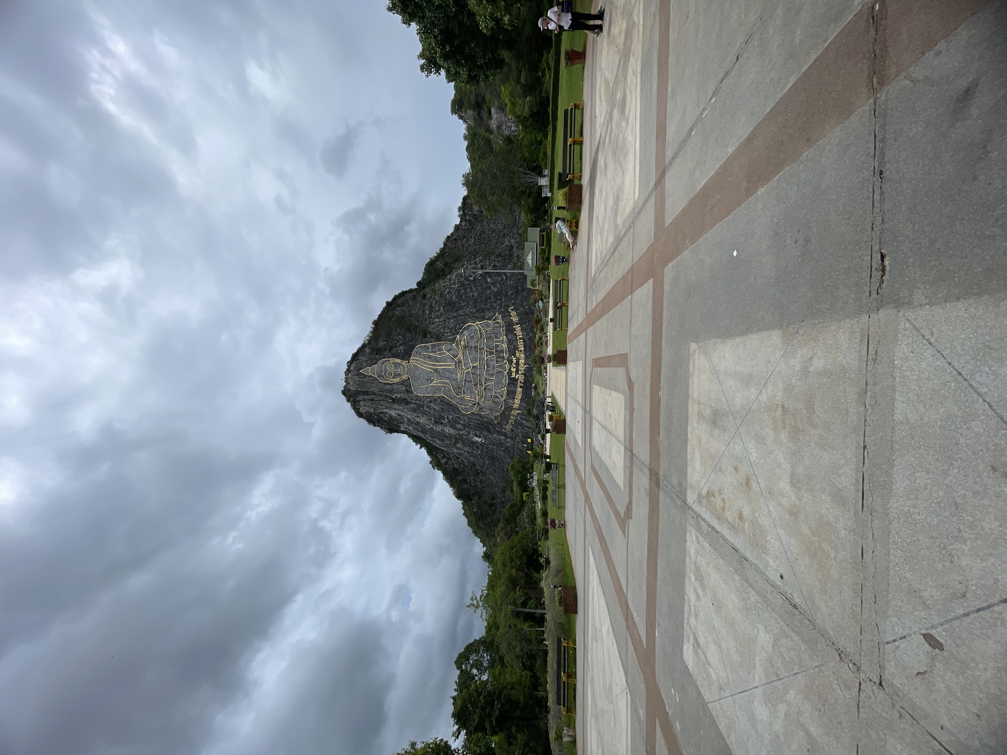 A large golden Buddha image is etched into the side of a rocky mountain under a cloudy sky. The Buddha is seated in a meditative pose. Below the mountain, there is a manicured lawn with several benches and trees. In the foreground, a paved plaza with geometric patterns leads towards the lawn. Two people are visible in the plaza; one is standing on the right side of the image, looking towards the mountain, and another is seated on a bench on the left side of the image, facing away from the camera. The area appears to be a public space, possibly a park or tourist attraction, located in Na Chom Thian, Thailand.