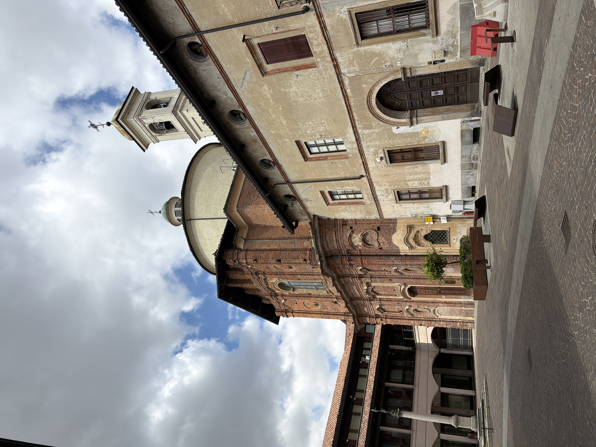 The image depicts a low-angle view of the Piazza Ducale in Vigevano, Italy. The architecture is characterized by the red brick and stucco façade of the Santa Maria delle Grazie church, which features a dome and a bell tower visible against a cloudy sky. Adjacent to the church, the image captures a section of the Palazzo Ducale, with windows, a large wooden door, and details of its ornate façade. The foreground shows the paved square with modern seating.