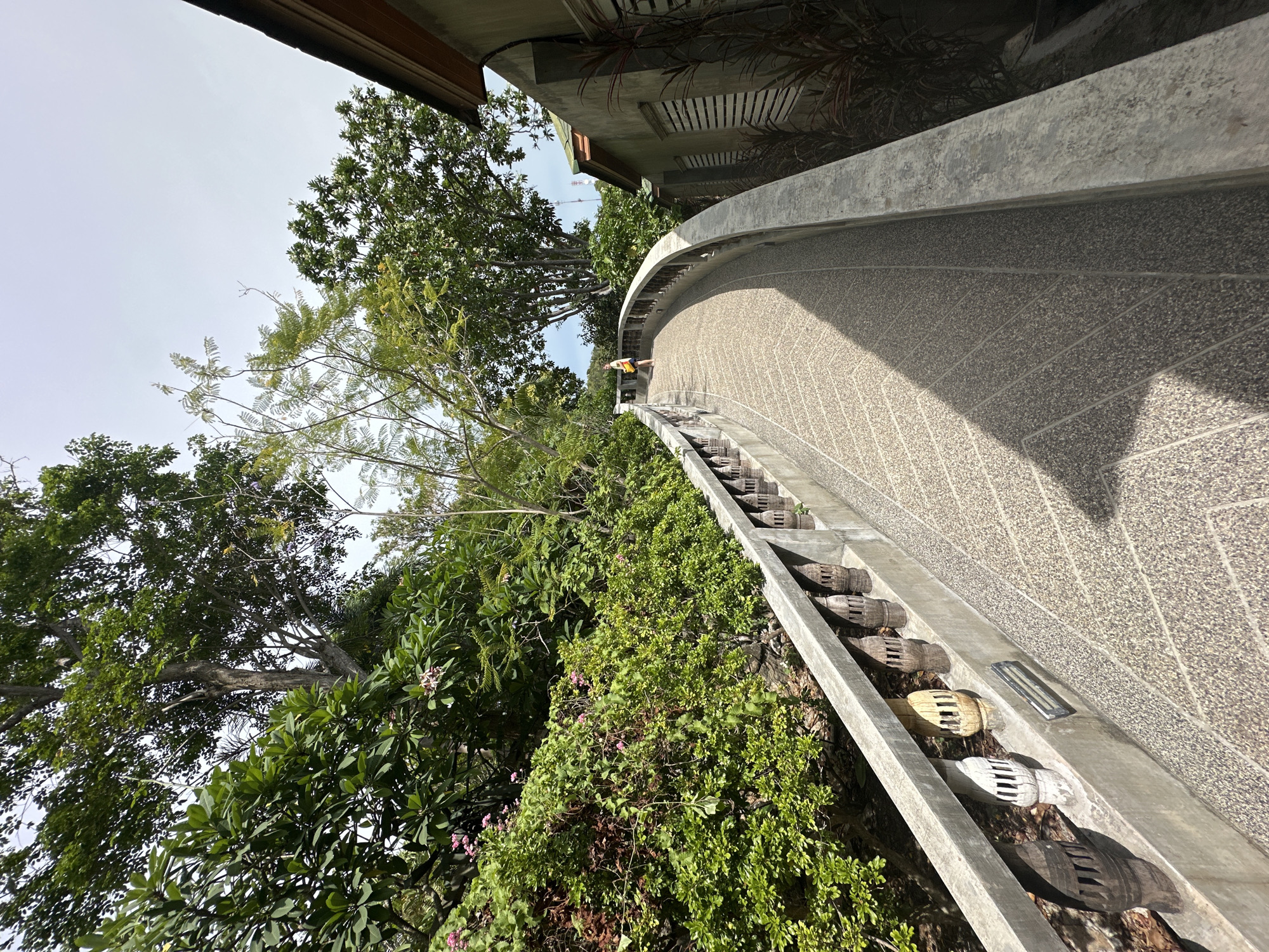 The image depicts a curved, elevated walkway made of concrete with a pebbled surface, situated in a lush, tropical environment. The walkway is bordered by a concrete railing with decorative, carved wooden posts, some of which appear weathered and varying in color from white to dark brown. On the left side of the walkway, dense green foliage, including bushes and trees with visible pink flowers, grows up a rocky incline. On the right side of the walkway, there is a building with a light green stucco exterior and a dark brown roof. The building has several windows with slatted shutters, and reddish-brown, spiky plants grow along its base. In the distance, a person wearing a yellow shirt and shorts is walking away from the camera on the walkway. The sky is overcast. The scene appears to be located at a tourist site or temple in Ko Pha Ngan, Thailand, likely in the Tambon Ko Pha Ngan area, given the vegetation and architectural style.