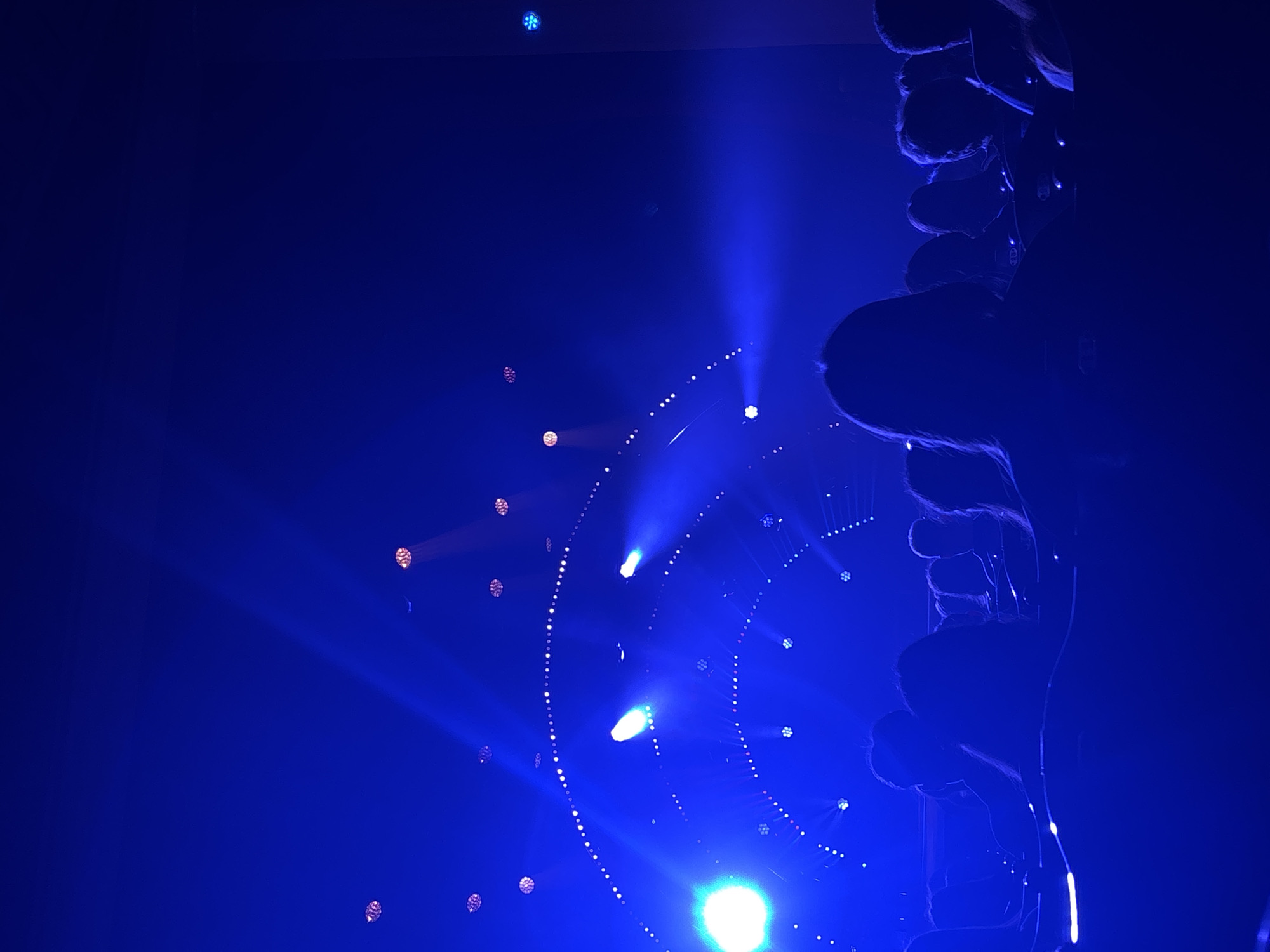 The image shows a nighttime scene illuminated by blue stage lighting. At right, there is a blurry silhouette of a group of people, possibly an audience or performers. A series of curved lines of small lights arc across the image, suggesting a decorative light display or architectural feature. Beams of blue light emanate from several points. The location is possibly Perm, Russia.