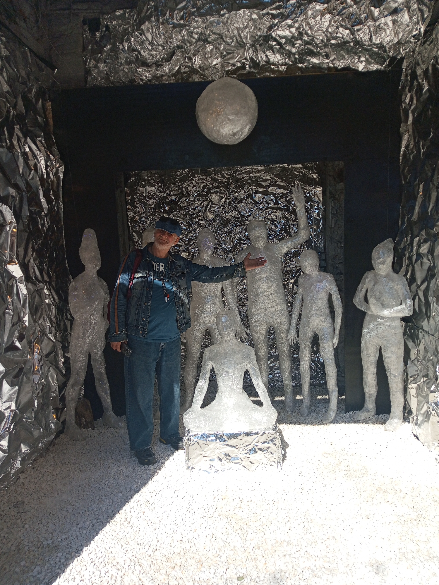 An interior space is decorated with crushed white rock on the floor, and aluminum foil covers the walls and ceiling. Six human-shaped sculptures made from clear plastic wrap or similar material are present; one is seated on a pedestal. A man in a denim jacket and cap stands in front of the figures, gesturing towards them with his right hand. A spherical object also covered in aluminum foil hangs from the ceiling. The location is stated as Saint Petersburg, Russia.