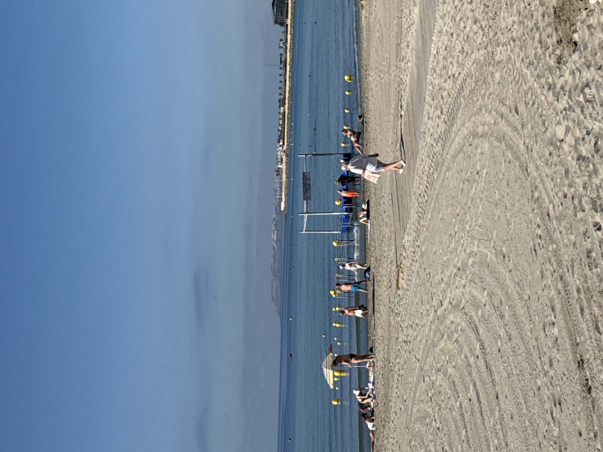 The image depicts a sunny day at a beach in Unknown, Unknown. The foreground is dominated by sand with tire tracks and footprints. A wooden walkway is visible on the right side of the image. Several people are present on the beach. In the mid-ground, a group of people are gathered near a colorful umbrella. Further back, near the water's edge, more individuals are visible, some standing and some walking. A man is entering the water, and a woman is walking away from it on the wooden pathway. Yellow buoys dot the calm blue water, which extends to the horizon. In the background, a distant coastline is visible with buildings and structures, suggesting a developed area beyond the beach. The sky is clear and blue.