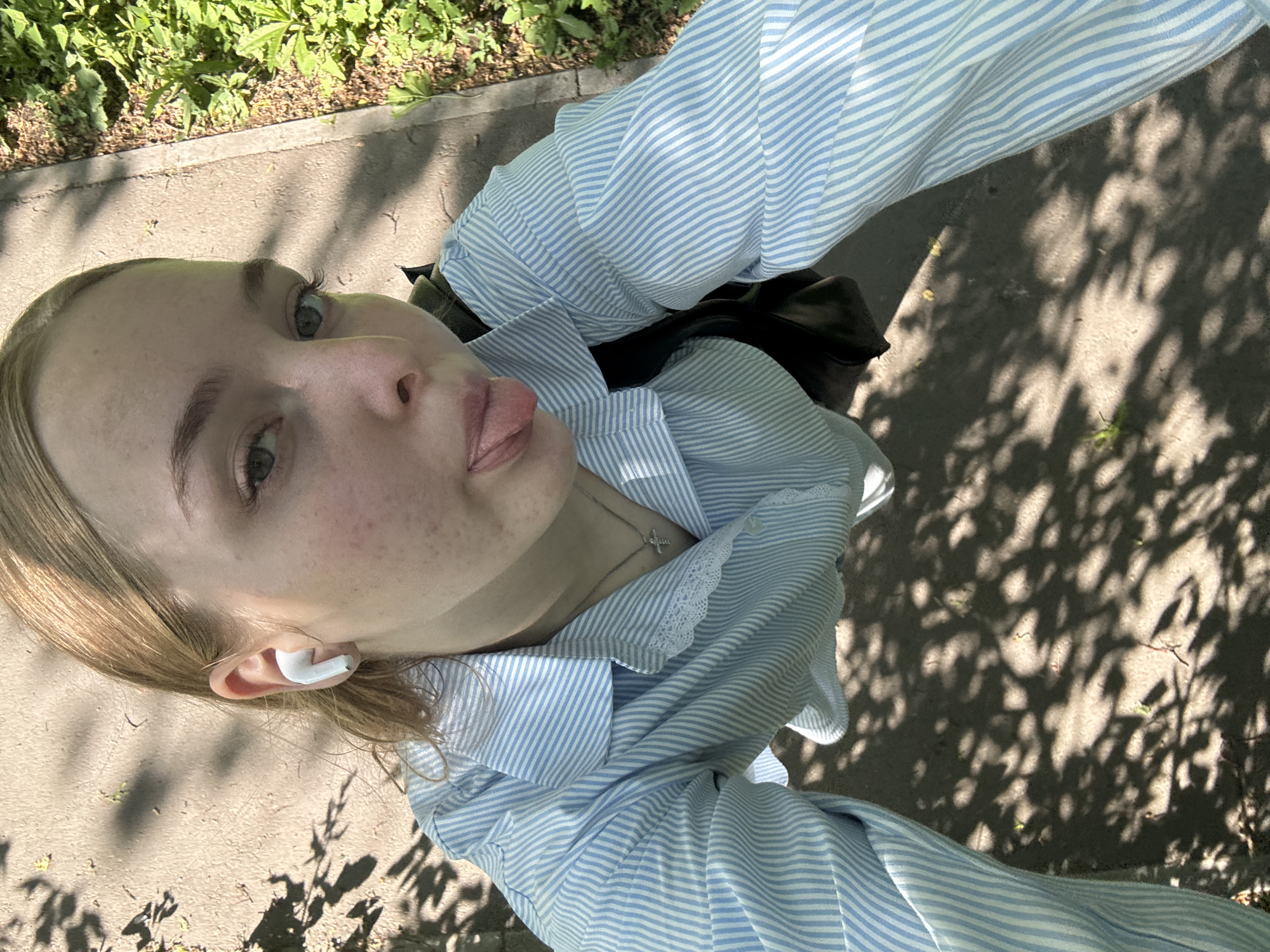 A woman is taking a selfie in Perm, Russia. She is wearing a light blue striped shirt and a silver necklace. She has a white earbud in her left ear. The background shows a shaded sidewalk with trees and greenery visible.