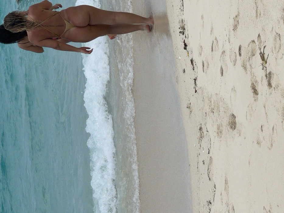 A beach scene in Zanzibar, Tanzania, depicts two individuals walking barefoot into the ocean. The foreground features light-colored sand with numerous footprints and scattered small pieces of natural debris. Gentle waves break on the shore, composed of clear, light blue-green water. The individual closer to the camera, a female, is viewed from behind, wearing a gold-colored thong-style one-piece swimsuit with thin crisscrossing straps across her back. Faint tan lines are visible on her skin. Ahead of her, partially obscured, a second individual, also appearing female, is visible from behind, wearing a dark-colored swimsuit. Both are progressing into the water, away from the viewer.