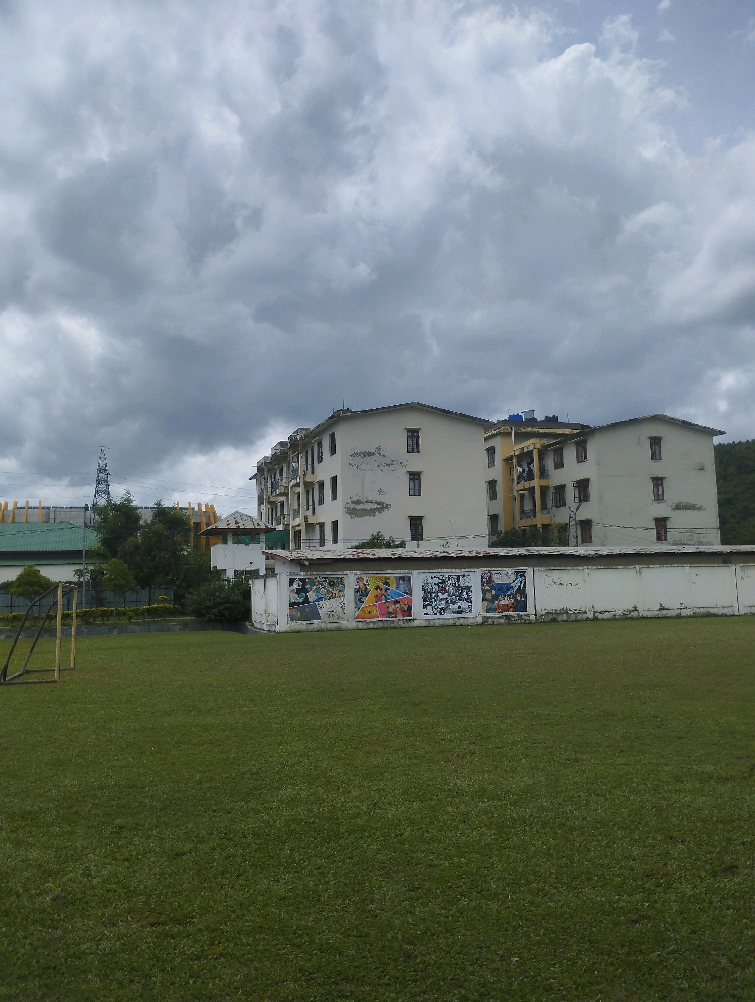 A ground-level view of a sports field with a goalpost on the left, in front of a long white wall decorated with colorful murals. Behind the wall are multi-story buildings with visible windows and balconies. The sky is overcast with dense clouds. The scene appears to be in Heingang, India.