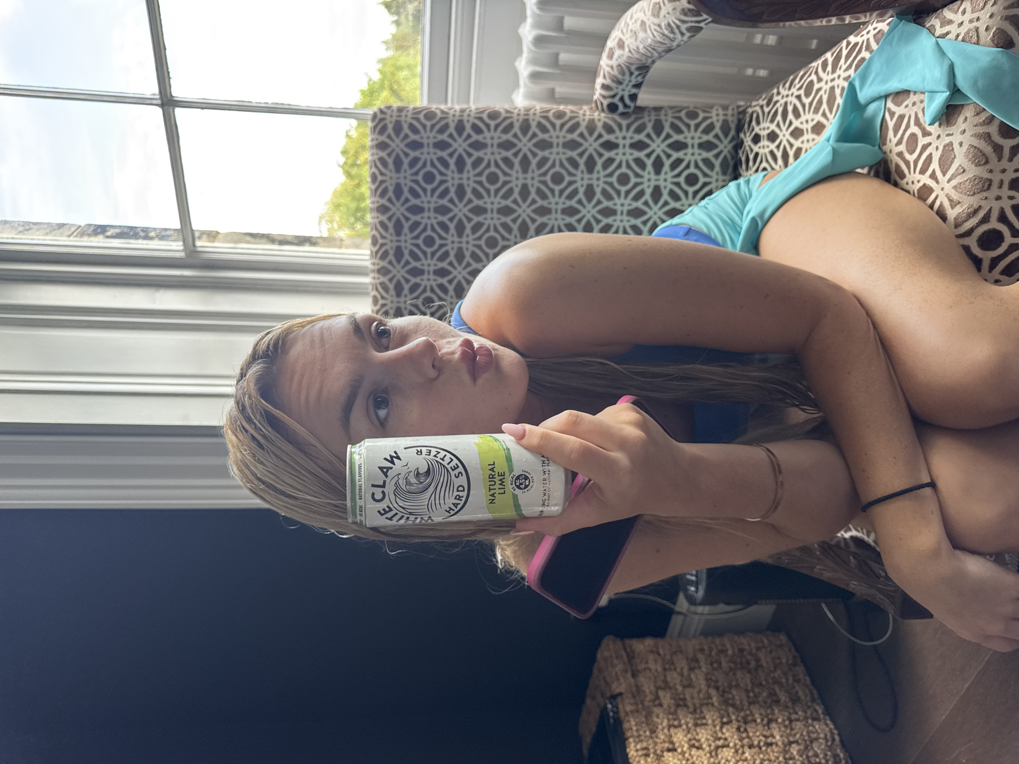 A young woman with blonde hair is seated in an armchair, holding a White Claw hard seltzer can in her right hand and a pink-cased smartphone in her left. She is wearing a blue dress. The armchair has a patterned upholstery. Behind her, a large window with multiple panes reveals greenery and a bright sky. To the left of the armchair, a woven basket and a dark surface are visible. The floor appears to be wooden. The lighting suggests it is daytime. The scene is indoors.