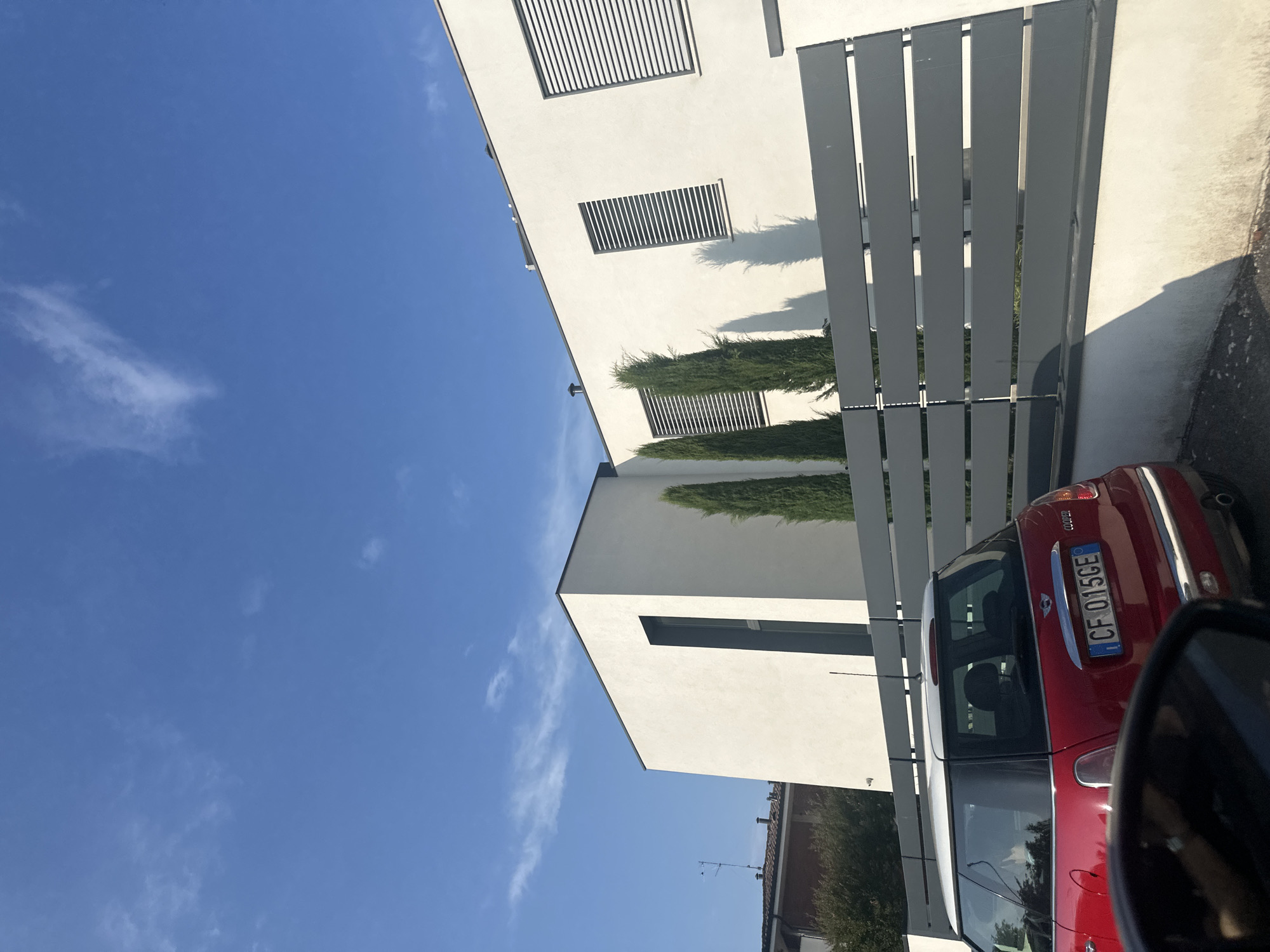 The image captures a sunny day scene outside a modern white house with a dark-trimmed roof, located in Chiari, Italy. A gray, horizontal-slatted fence separates the property from the street. Three tall, slender cypress trees stand behind the fence, casting shadows on the white wall of the house. In the foreground, a red Mini Cooper car with a license plate reading "CF 015GE" is visible, parked in front of the fence. The sky above is a clear, bright blue with a few wispy white clouds.