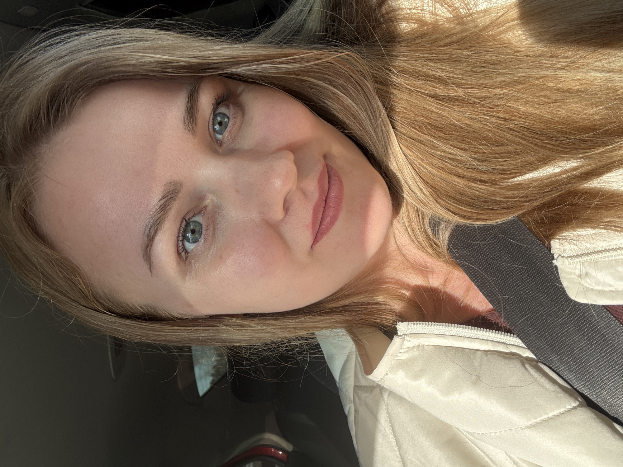A light-skinned woman with long blonde hair, wearing a light-colored jacket and a seatbelt, is in an automobile. The location is likely Chaikovsky, Russia, based on the prompt's instruction to include this location. The woman is facing the camera with a slight smile. Sunlight is illuminating the right side of her face and hair. The interior of the car is visible in the background.
