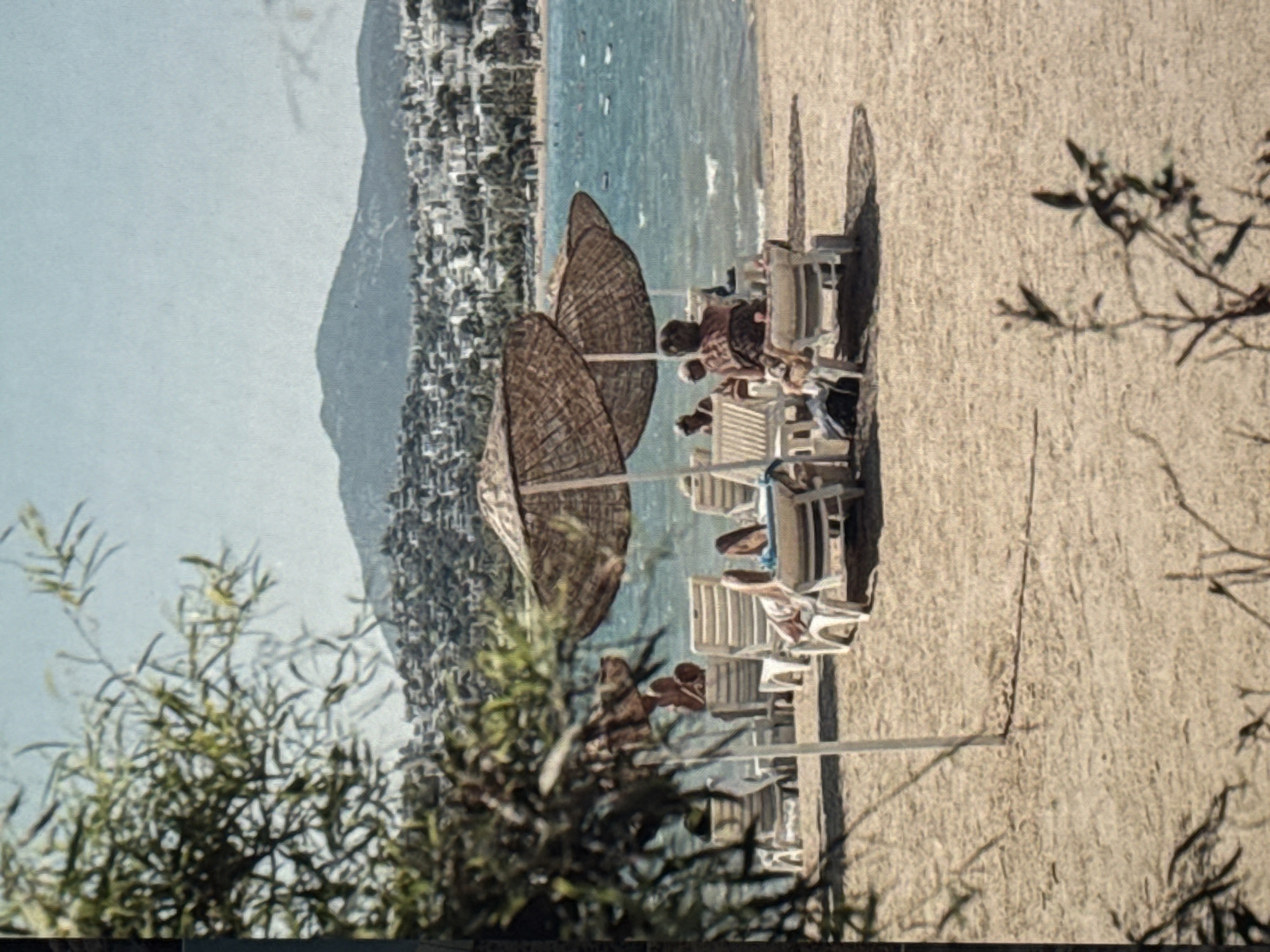 This is a vertical, slightly grainy, medium-shot photograph of a beach scene in Eyüpsultan, Turkey. The sun is bright, casting long shadows, suggesting it is late afternoon. The sea is a clear, turquoise blue, with a few boats visible on the horizon. Behind the beach, a densely populated hillside covered in buildings ascends, characteristic of a coastal town.
On the sand, there are several white plastic beach chairs, some occupied by people lounging and relaxing. Two large, woven straw beach umbrellas provide shade. One umbrella is prominently in the foreground, and another is further back. The people are casually dressed, enjoying the warm weather. One person in a chair appears to be reading. The overall mood is one of relaxation and leisure on a summer day. The sand is light brown and appears smooth. In the lower left corner, out-of-focus green foliage is visible, indicating the presence of vegetation near the beach. There is no visible text in the image.