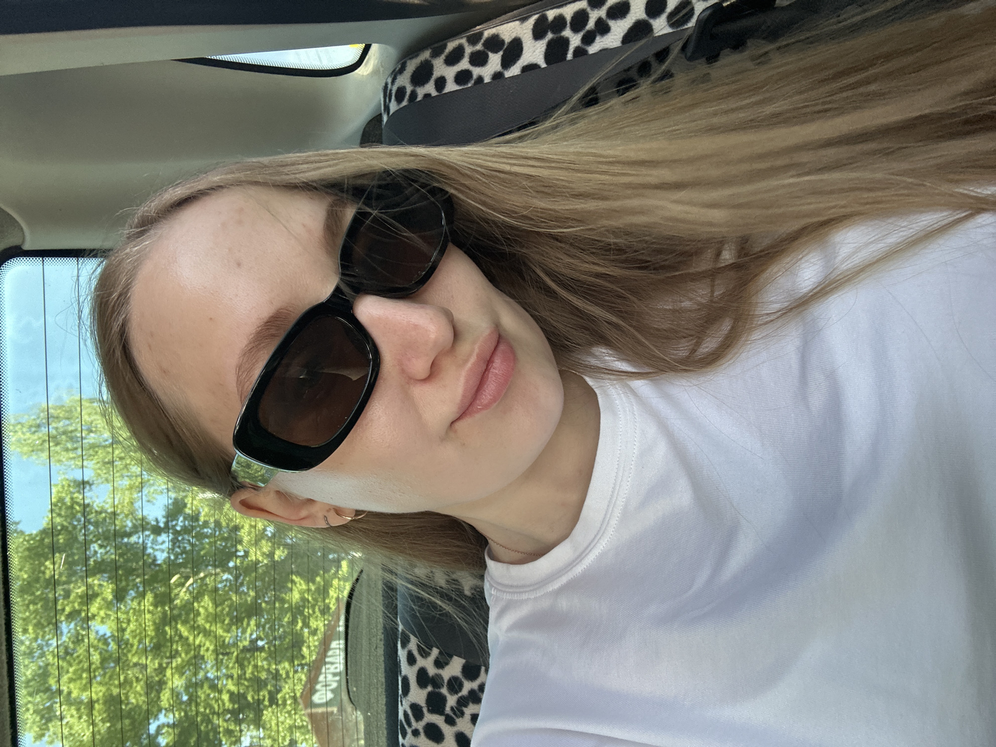 A young woman with blonde hair and sunglasses is in a car, possibly as a passenger. She is wearing a white t-shirt and appears to be taking a selfie. The car's interior has a black and white patterned headrest. Through the car window, trees and the corner of a building are visible, with Cyrillic script on a sign, suggesting a location in a Russian-speaking area. The building and sign details corroborate the location being in Chaikovsky, Russia.
