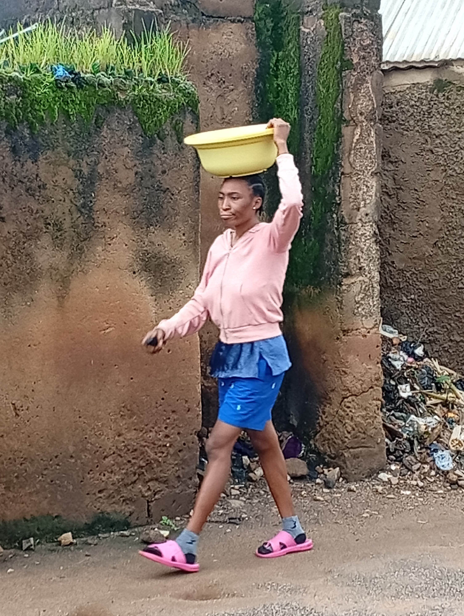 A young woman is captured walking with a yellow basin balanced on her head, against a backdrop of a textured, earth-toned wall. She is wearing a light pink hooded jacket over a dark blue skirt and bright blue shorts. Her feet are adorned with pink sandals and grey socks. The wall, which appears weathered and possibly made of mud or concrete, has patches of green moss and vegetation growing along its top edge. To the right of the woman, the scene transitions to a pile of discarded trash, including plastic bags and assorted debris, situated against a rougher textured wall. The ground in the foreground is a dusty, unpaved surface. This scene is likely located in Jos, Nigeria, given the prevalent architectural style and the surrounding environmental elements.