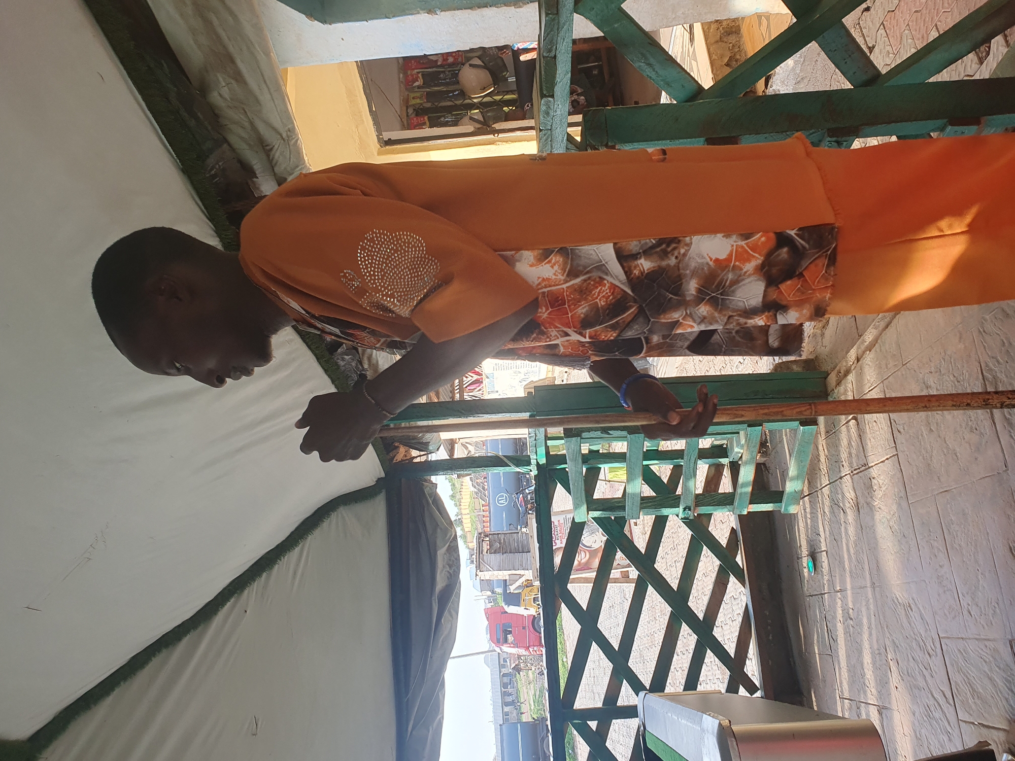 A person is standing outdoors under a white tent, holding a wooden pole. The person is wearing an orange garment with a patterned fabric insert and decorative rhinestones on the sleeve. In the background, a red truck and other vehicles are visible on a road, along with buildings and a green railing. The scene appears to be in Lokoja, Nigeria.