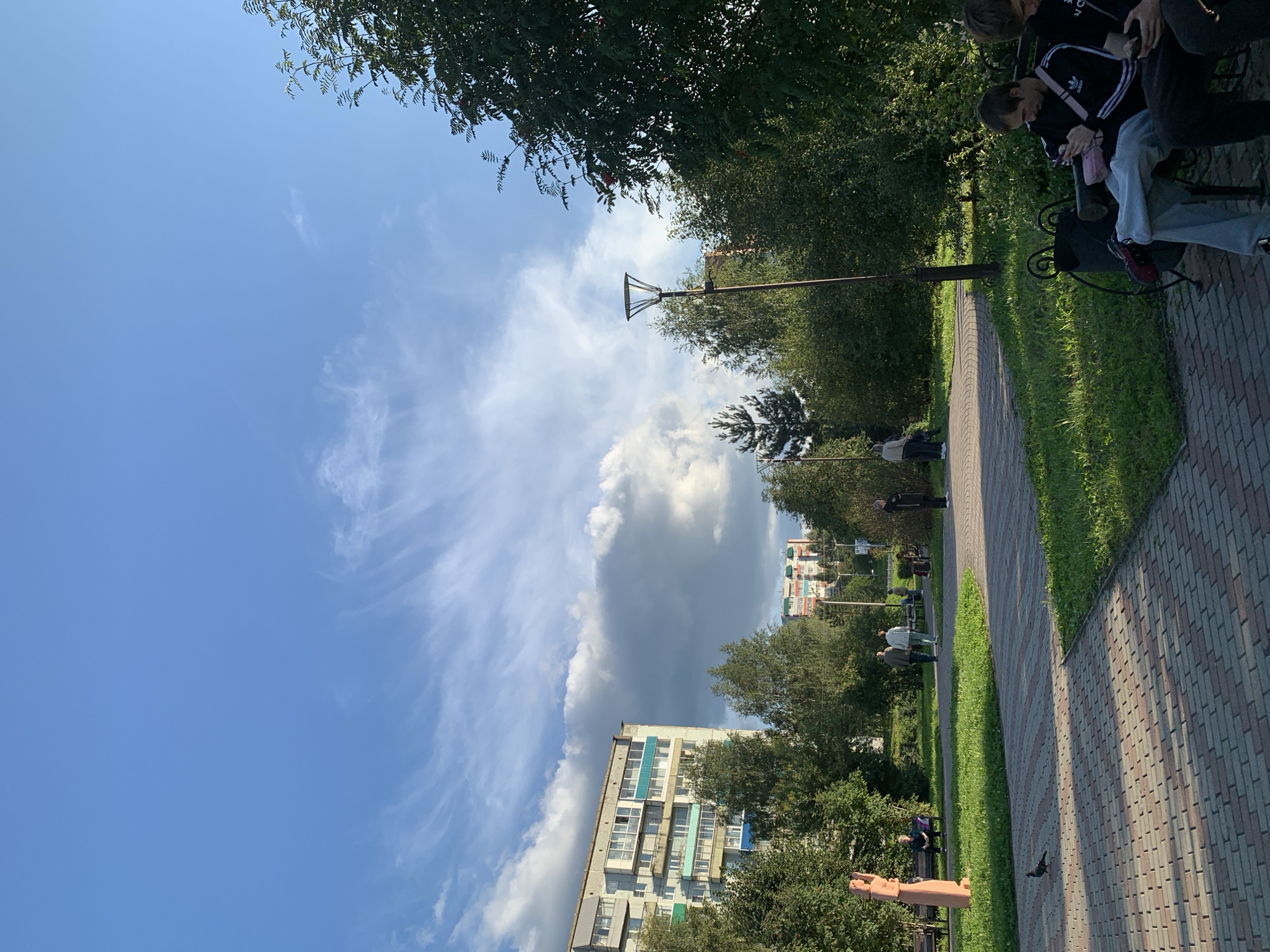 This image captures a park scene in Prokopyevsk, Russia. In the foreground, a brick-patterned pathway leads into a grassy area. To the left, a light brown, abstract sculpture stands near a park bench where a person is seated. Further back, several individuals are walking along another pathway. On the left side of the image, a multi-story apartment building with balconies is visible, partially obscured by trees. Across from it, on the right, a tall street lamp stands amidst lush green trees. In the distance, more apartment buildings can be seen. Several people are seated on a bench in the lower right corner, two of whom are looking at their phones. The sky above is a vibrant blue with wispy white clouds and darker storm clouds gathering in the distance. A pigeon is visible on the pathway in the lower left.