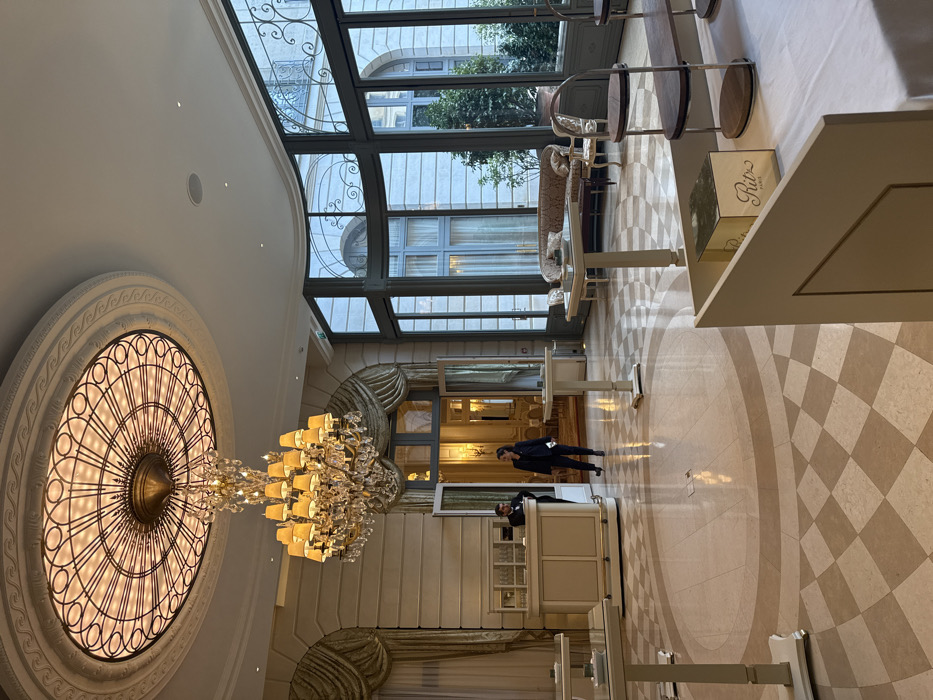 The image depicts the interior of a luxurious building, possibly a hotel. A large, ornate chandelier hangs from the ceiling next to a decorative circular window. Natural light floods in through a glass ceiling, illuminating a checkered floor. A concierge station is visible with two people, and tables with chairs line the area. The overall impression is one of opulence and grandeur.